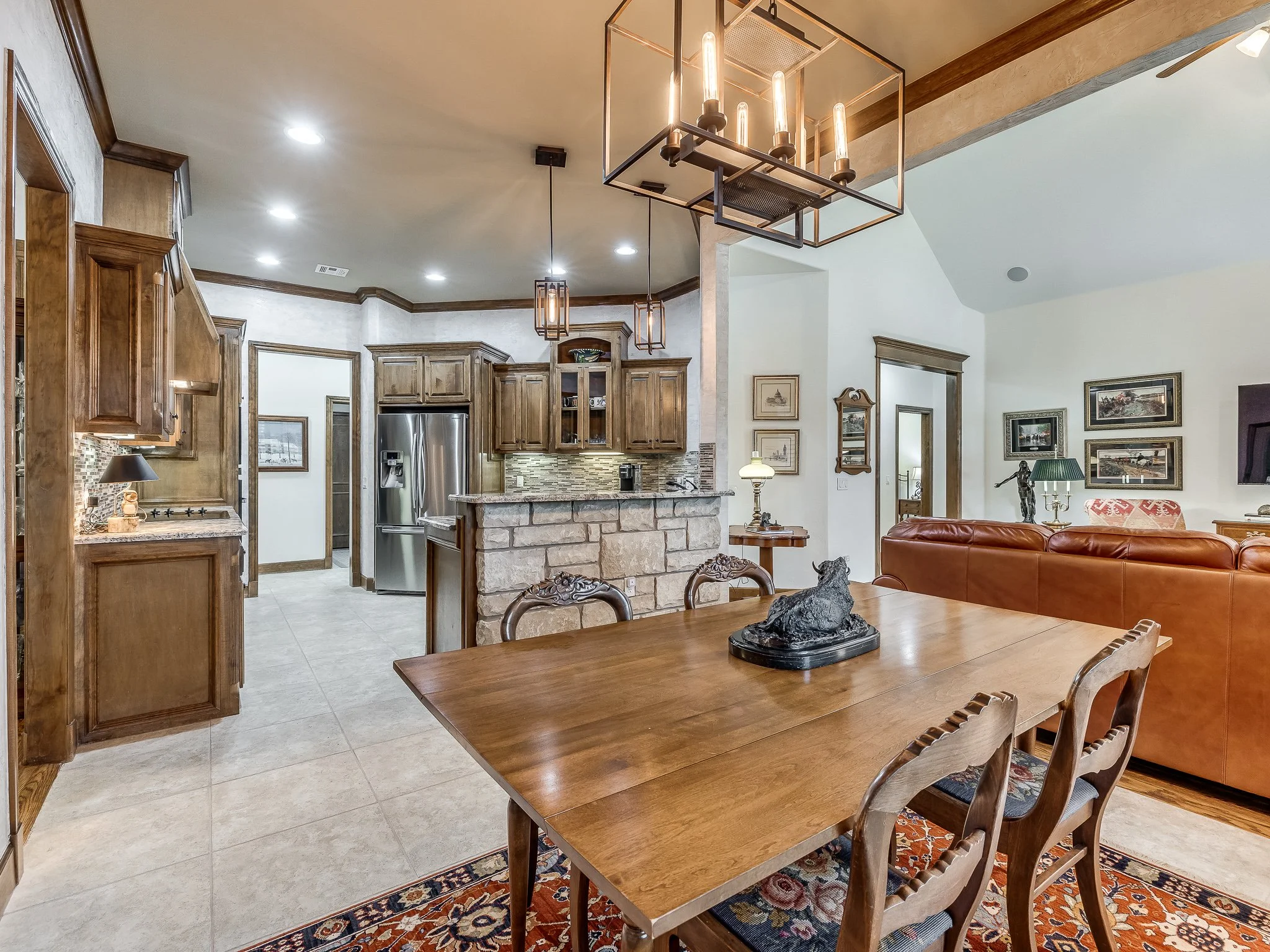 A spacious open-concept living area with a dining table in the foreground, a kitchen with wooden cabinets, stainless steel refrigerator, and stone accents in the background, and a cozy seating area with a leather sofa and decorative artwork on the walls.