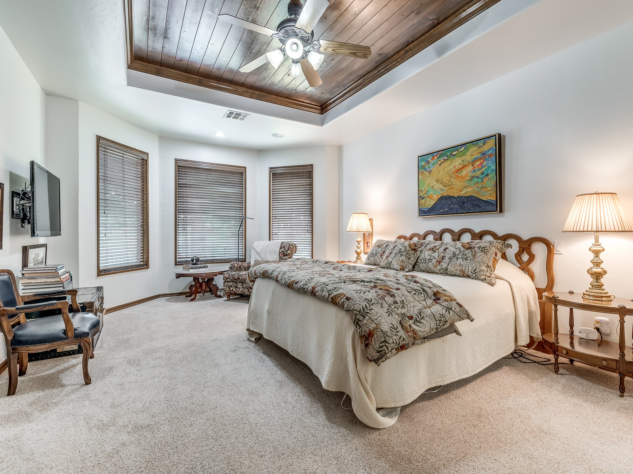 A spacious bedroom with a large bed, wooden headboard, floral bedding, and two nightstands with lamps. There is a ceiling fan with lights, three windows with blinds, a wall-mounted TV, and a small sitting area with chairs and a table. A colorful landscape painting hangs above the bed.