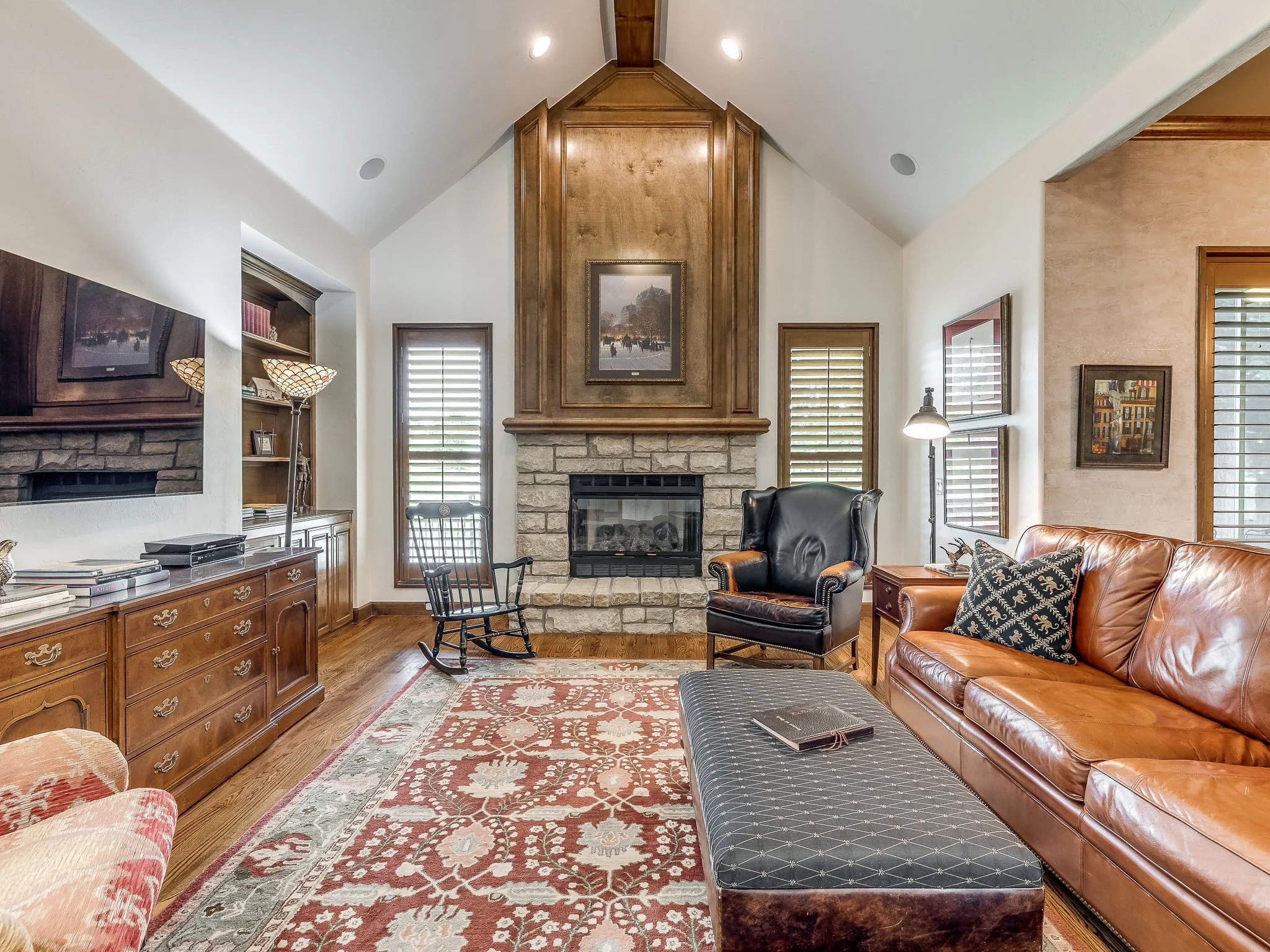 Living room with a stone fireplace, wooden mantle, brown leather sofa, black armchair, area rug, windows with blinds, and wall art.