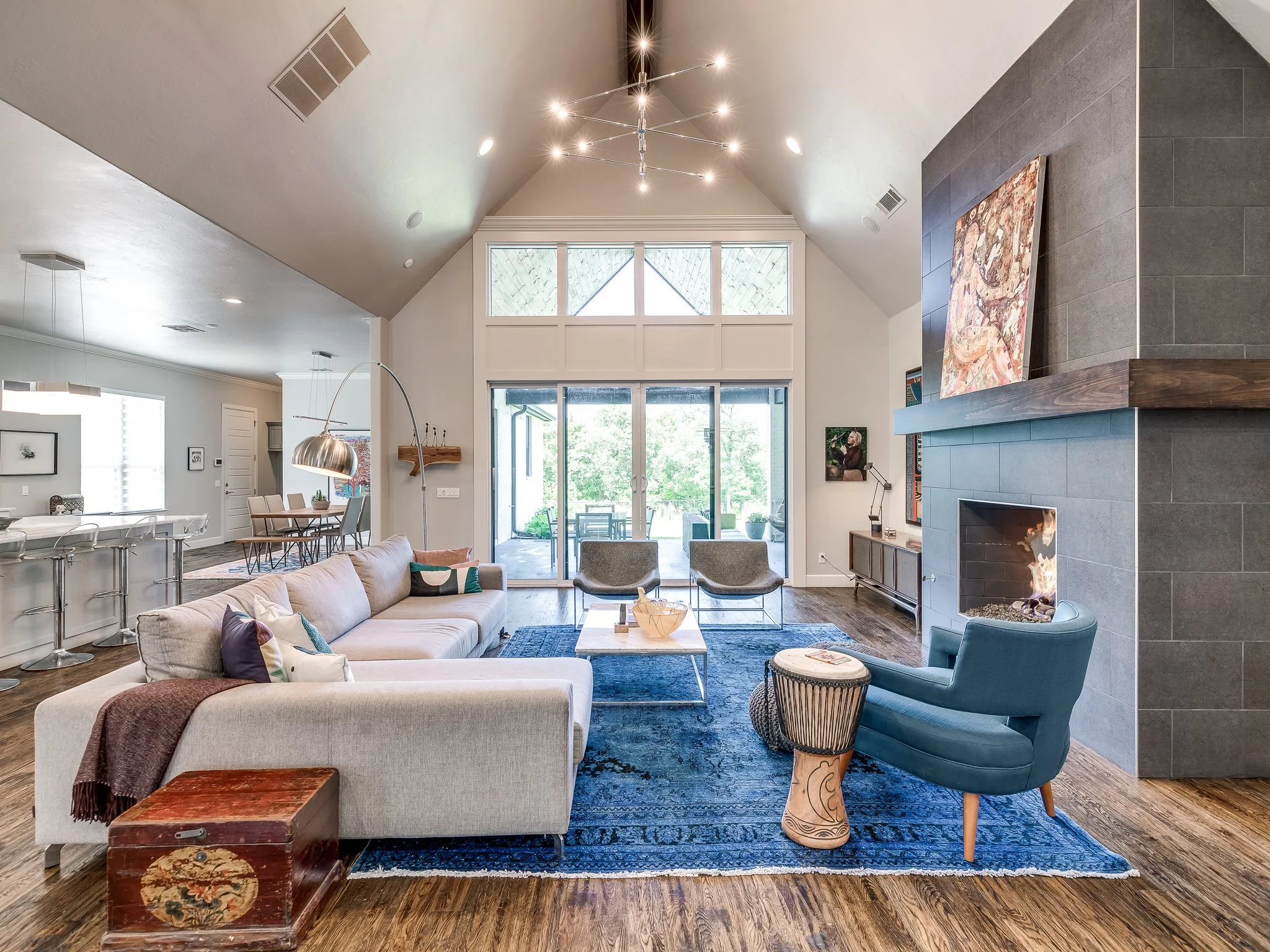Living room with high vaulted ceiling, modern chandelier, large window with sliding glass doors leading to outdoor patio, fireplace with gray tiles, blue armchair, beige sofa, wooden chest, and decorative rugs.