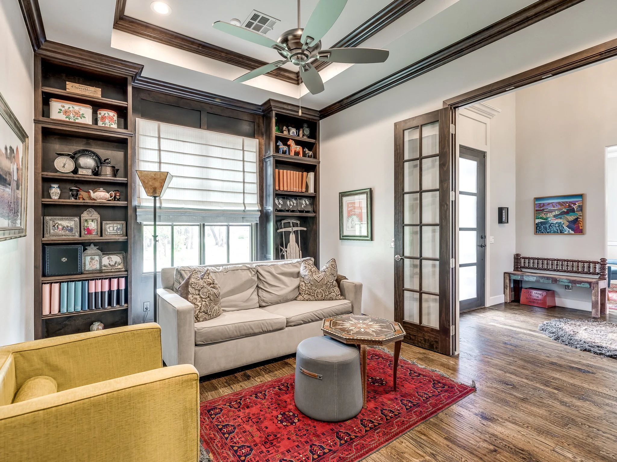 A cozy living room with a white sofa, an armchair, a wooden side table, and a red patterned rug. Dark wood built-in shelves filled with decor and books frame a window with a white roller shade. An open glass-paneled door leads to another room with a bench and colorful artwork, and hardwood floors are throughout.