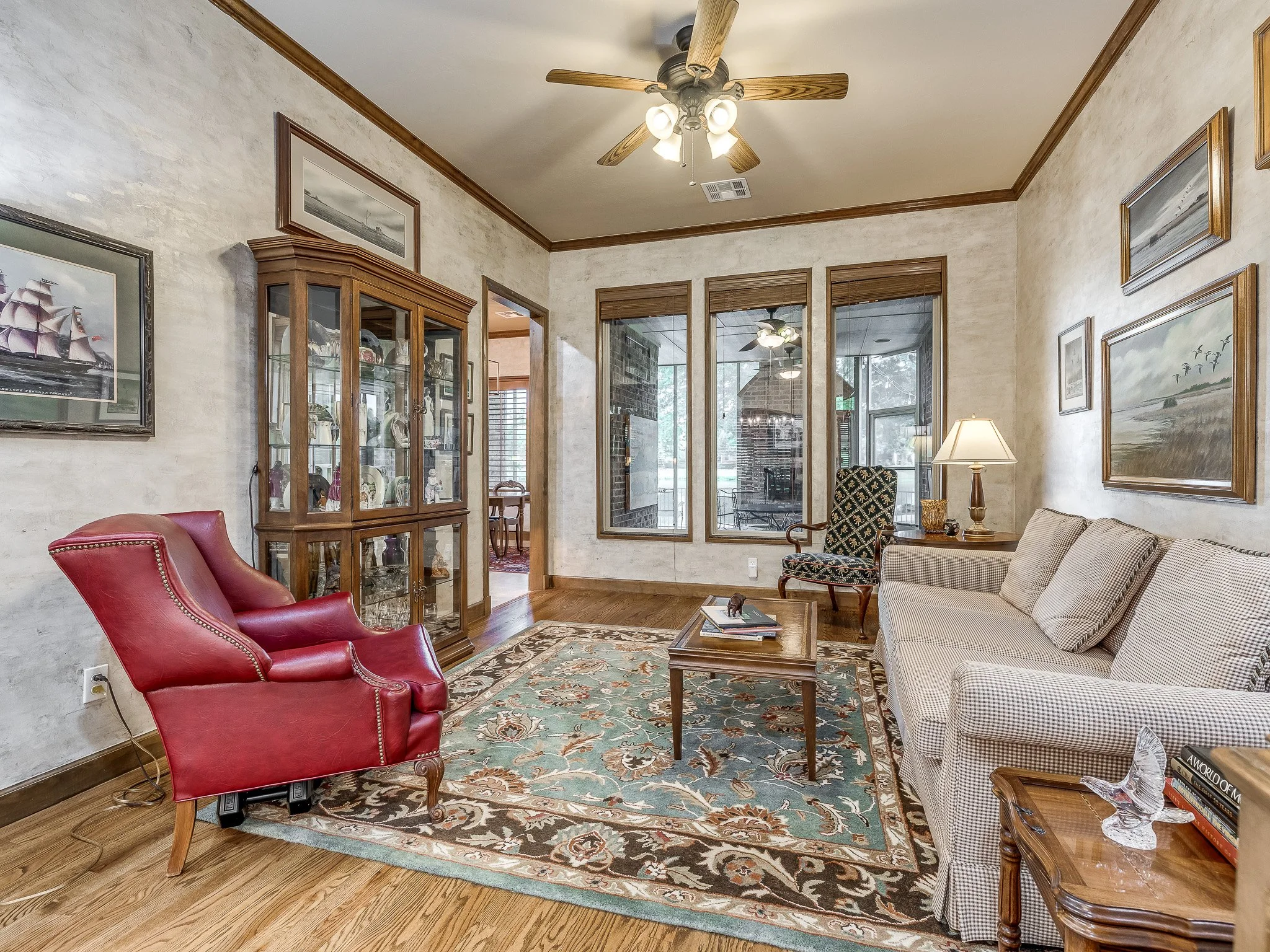 Living room with a red armchair, beige sofa, wooden coffee table, patterned rug, display cabinet with glass doors, armchair with a patterned fabric, table lamp, and framed artwork on walls, with large windows and a ceiling fan.