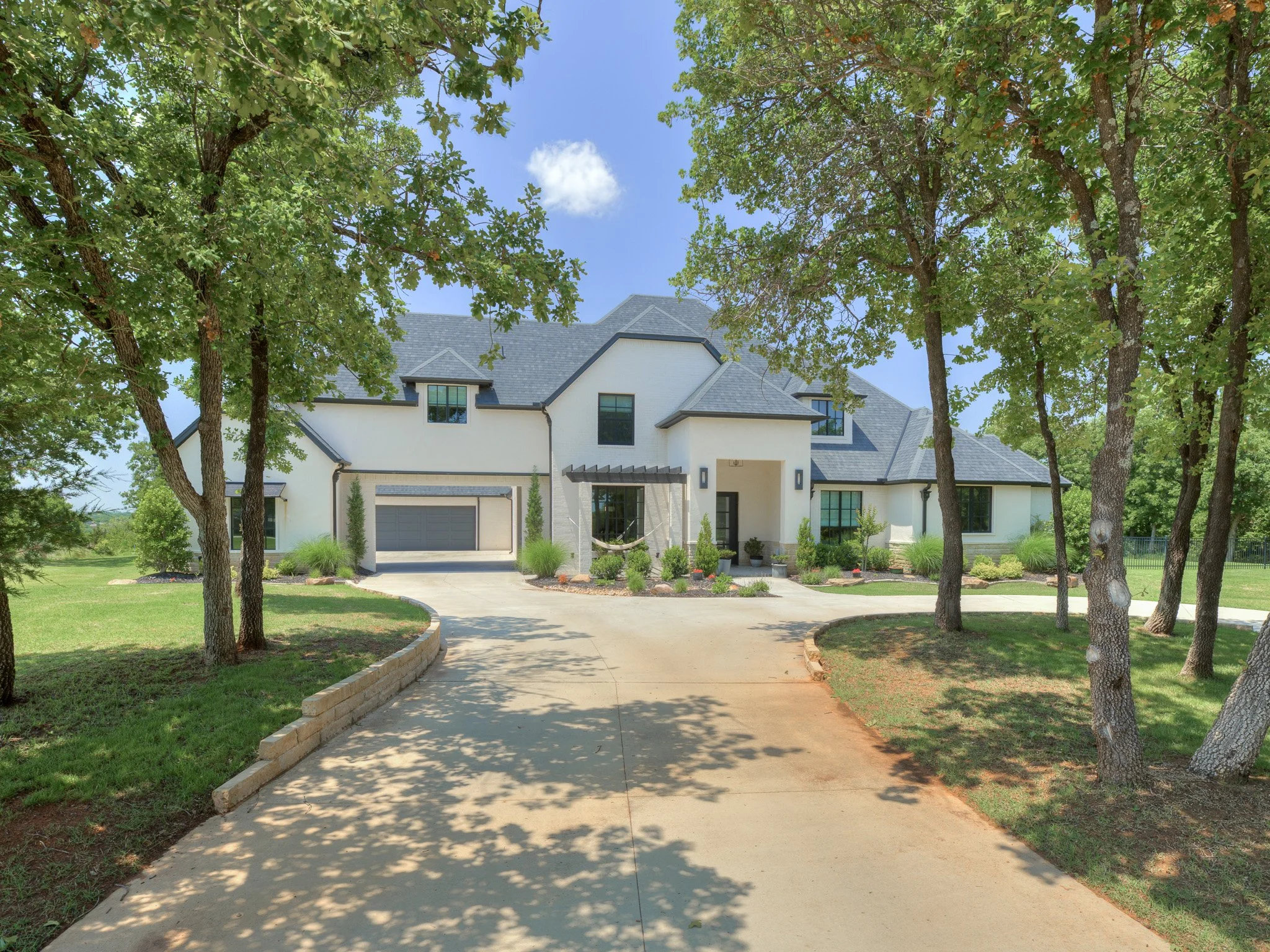 Modern two-story white house with gray roof, surrounded by trees and landscaped yard, with a curved driveway leading to the garage on a sunny day.