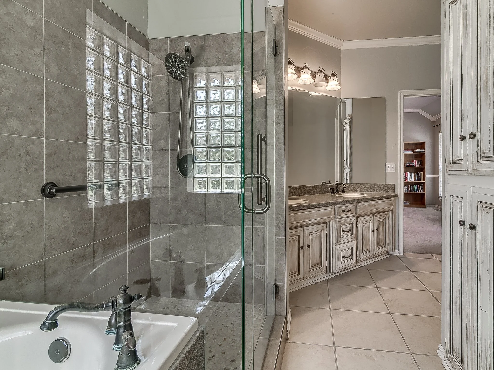 Bathroom view with a glass-enclosed shower, a double vanity with a mirror and four light fixtures above, beige cabinets, and a glimpse into an adjacent room with a bookshelf.