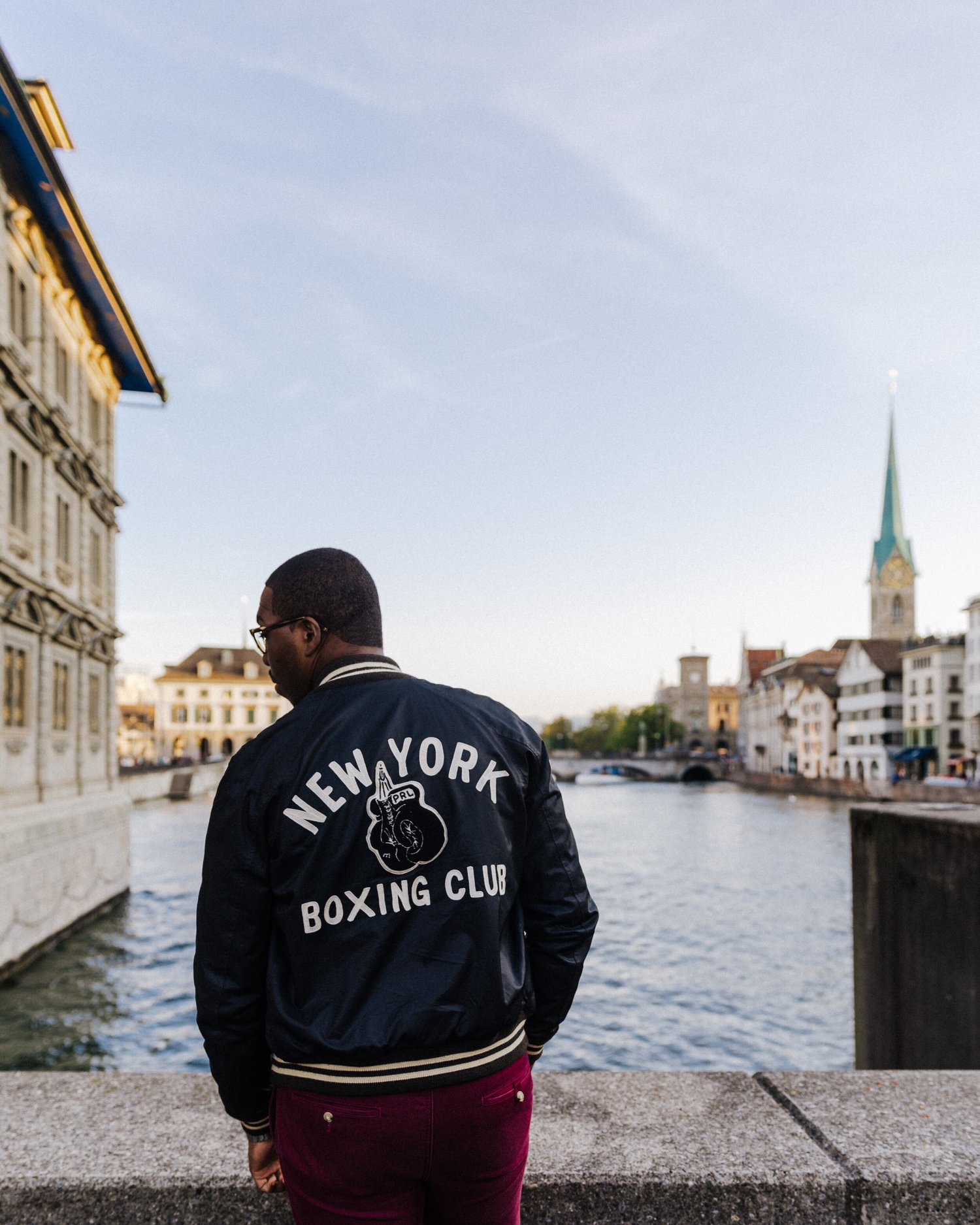 Maurice Jackson wearing a New York Boxing Club bomber jacket beside the river in Zurich