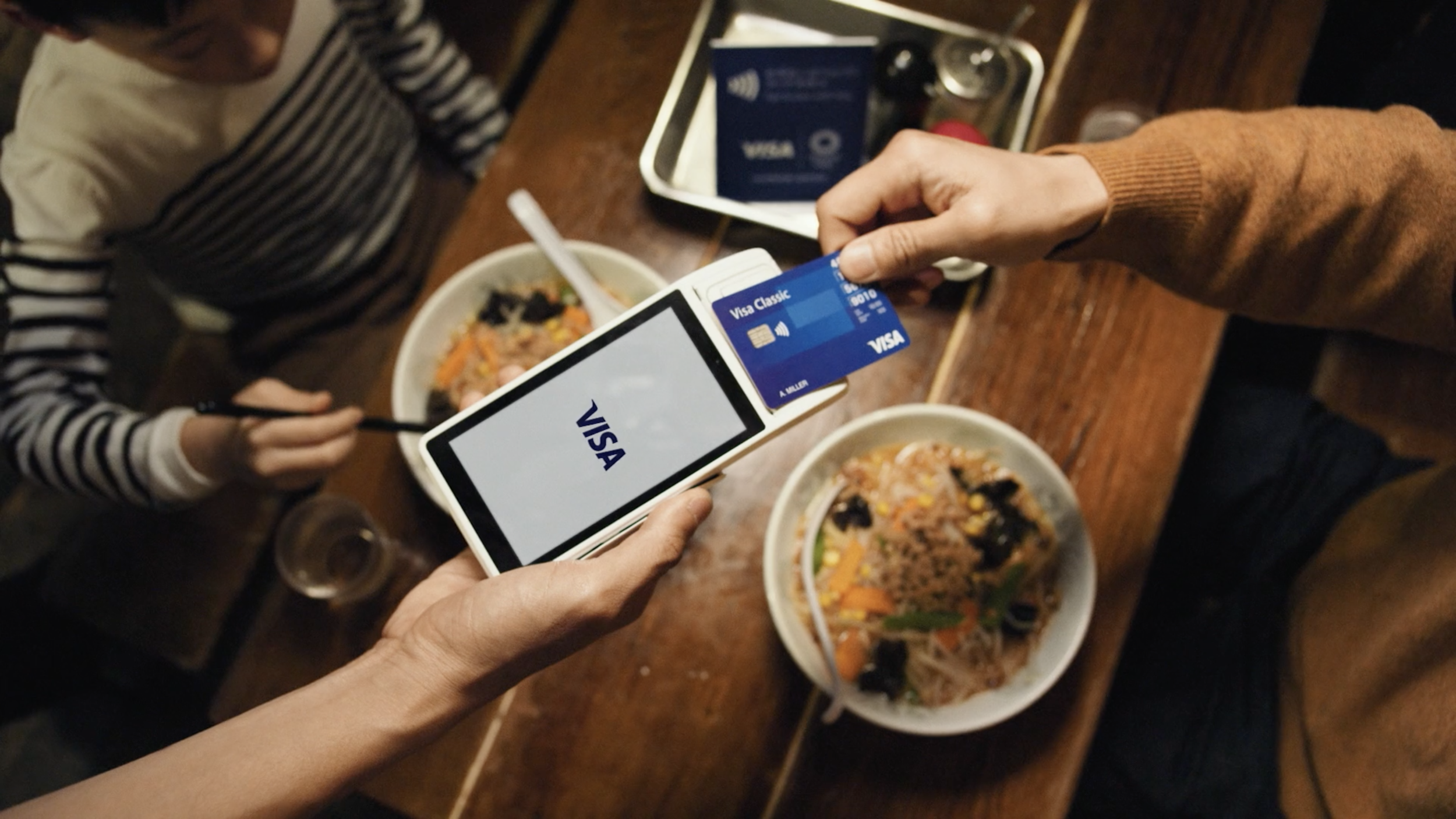 People at a restaurant using Visa credit cards to pay for bowls of Asian cuisine on a wooden table.