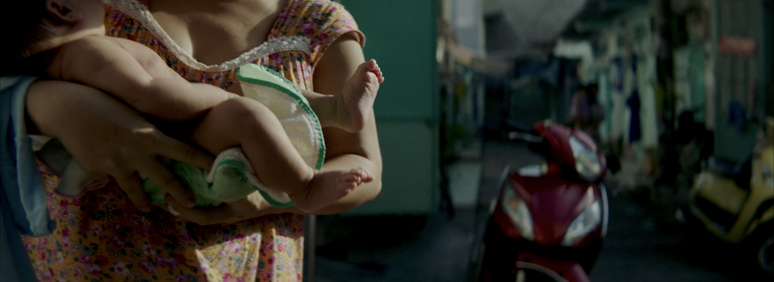 Person holding a baby in their arms in a busy outdoor setting with blurred background including a red motorcycle and people.