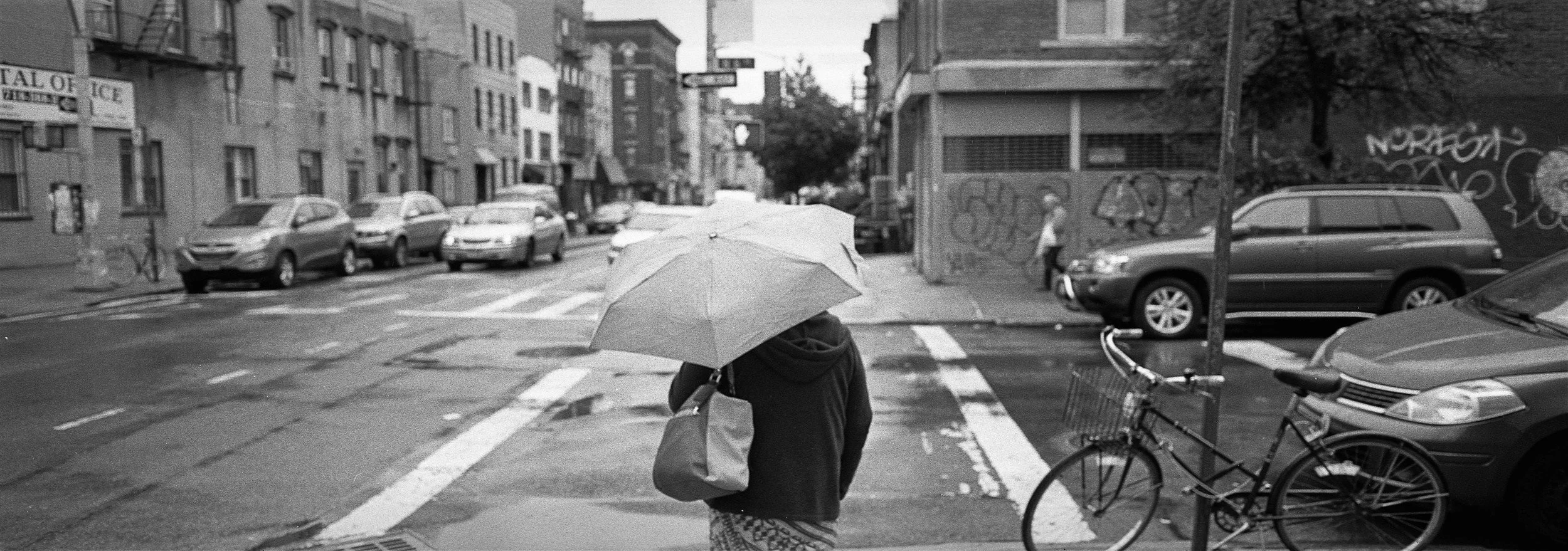 A person walking on a city street holding an umbrella, with parked cars, graffiti-covered walls, and buildings in the background.