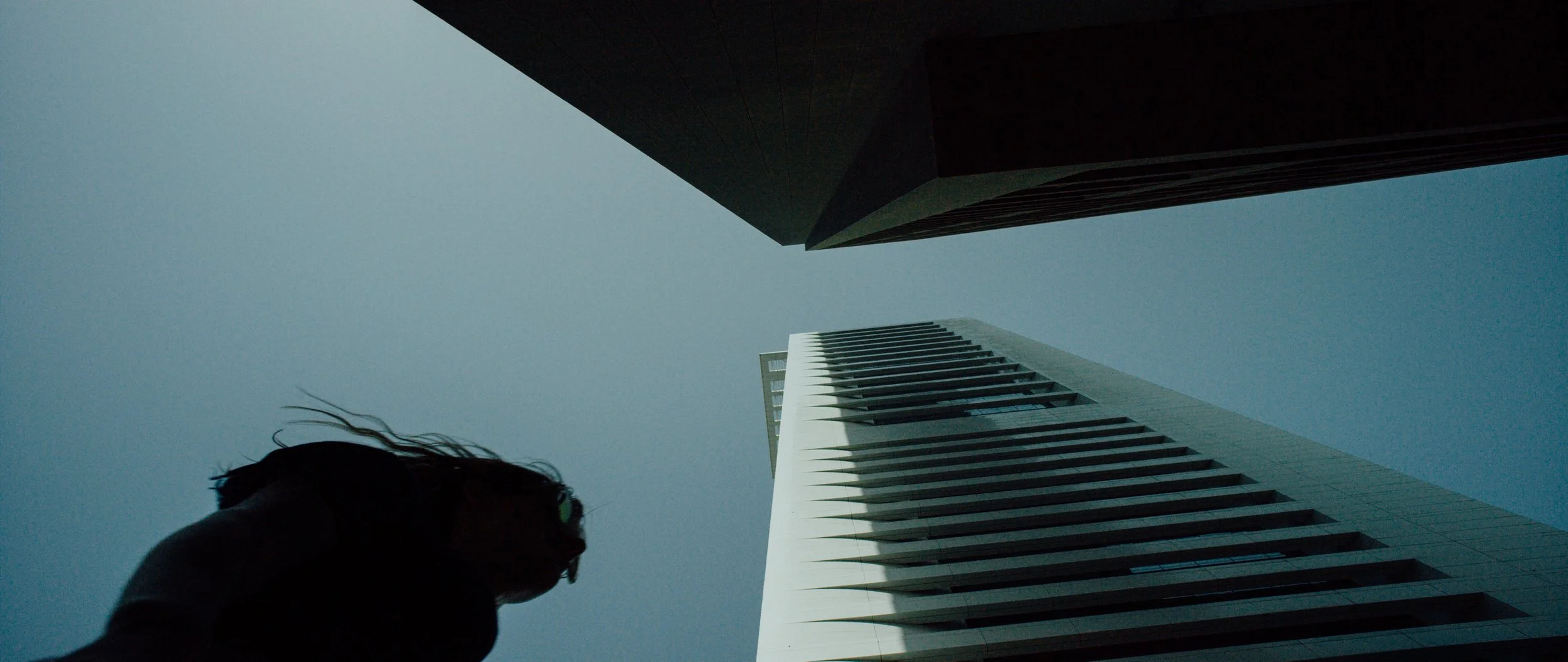 Low-angle view of two tall modern buildings against a blue sky, with a person's silhouette in the foreground.