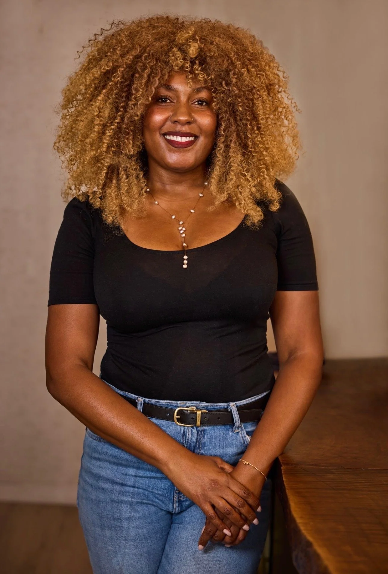A woman with curly blonde hair, wearing a black top, blue jeans, and jewelry, standing indoors against a neutral-colored background.