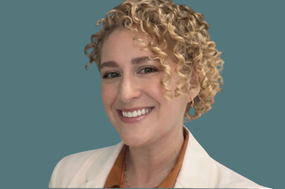 Portrait of a woman with short, curly blonde hair, wearing a white blazer and a brown blouse, smiling against a teal background.