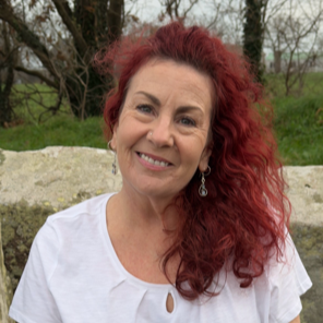 A woman with curly red hair smiling outdoors, wearing a white top and earrings, with trees and grass in the background.