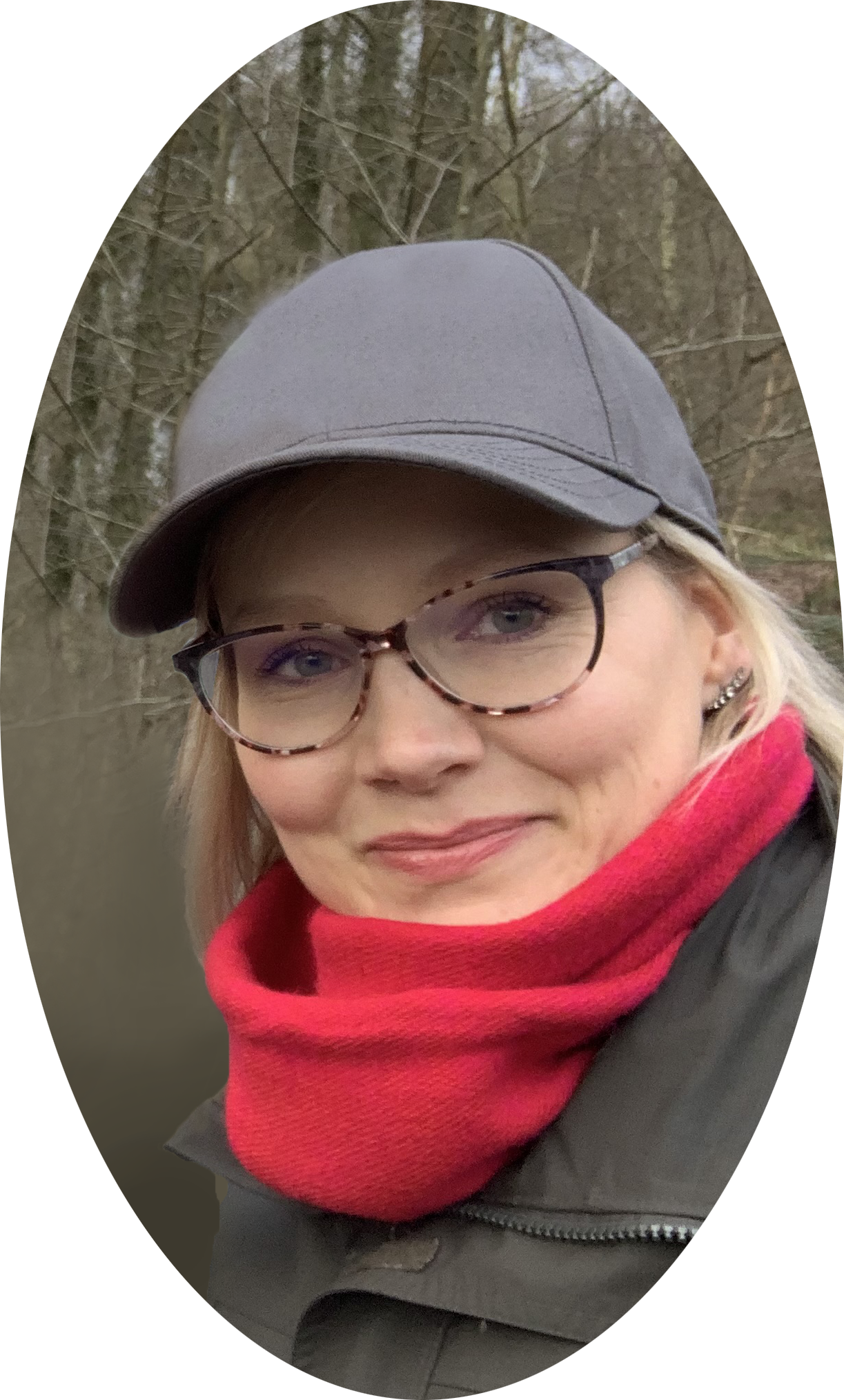 Een glimlachende vrouw met blond haar, bruin tortie montuurbrillen, grijze cap, rode sjaal en een donkere jas, in een natuuromgeving met bomen en een rivier.