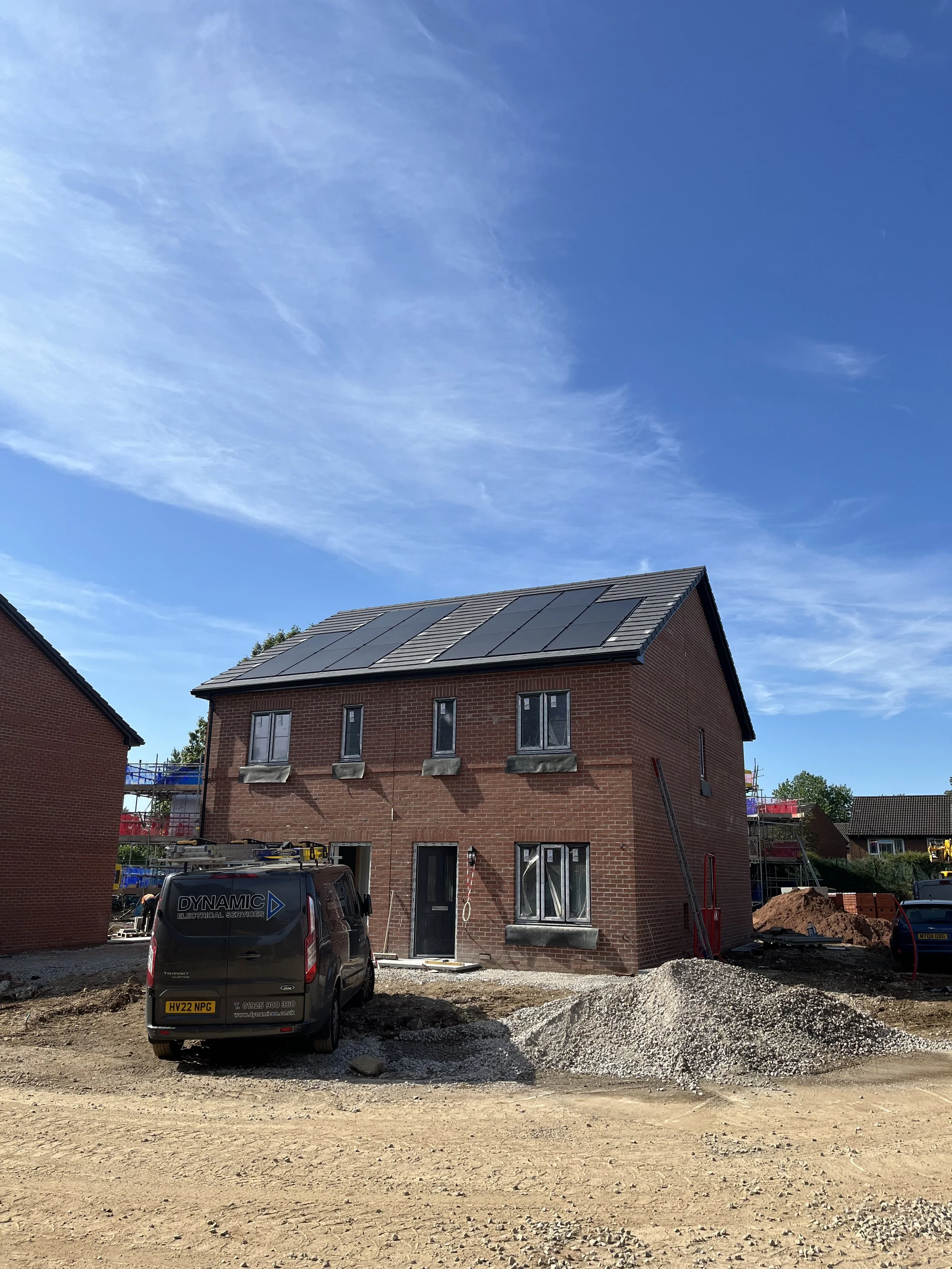 A house under construction with solar panels on the roof, surrounded by construction materials, tools, and a work van, on a sunny day with a blue sky.