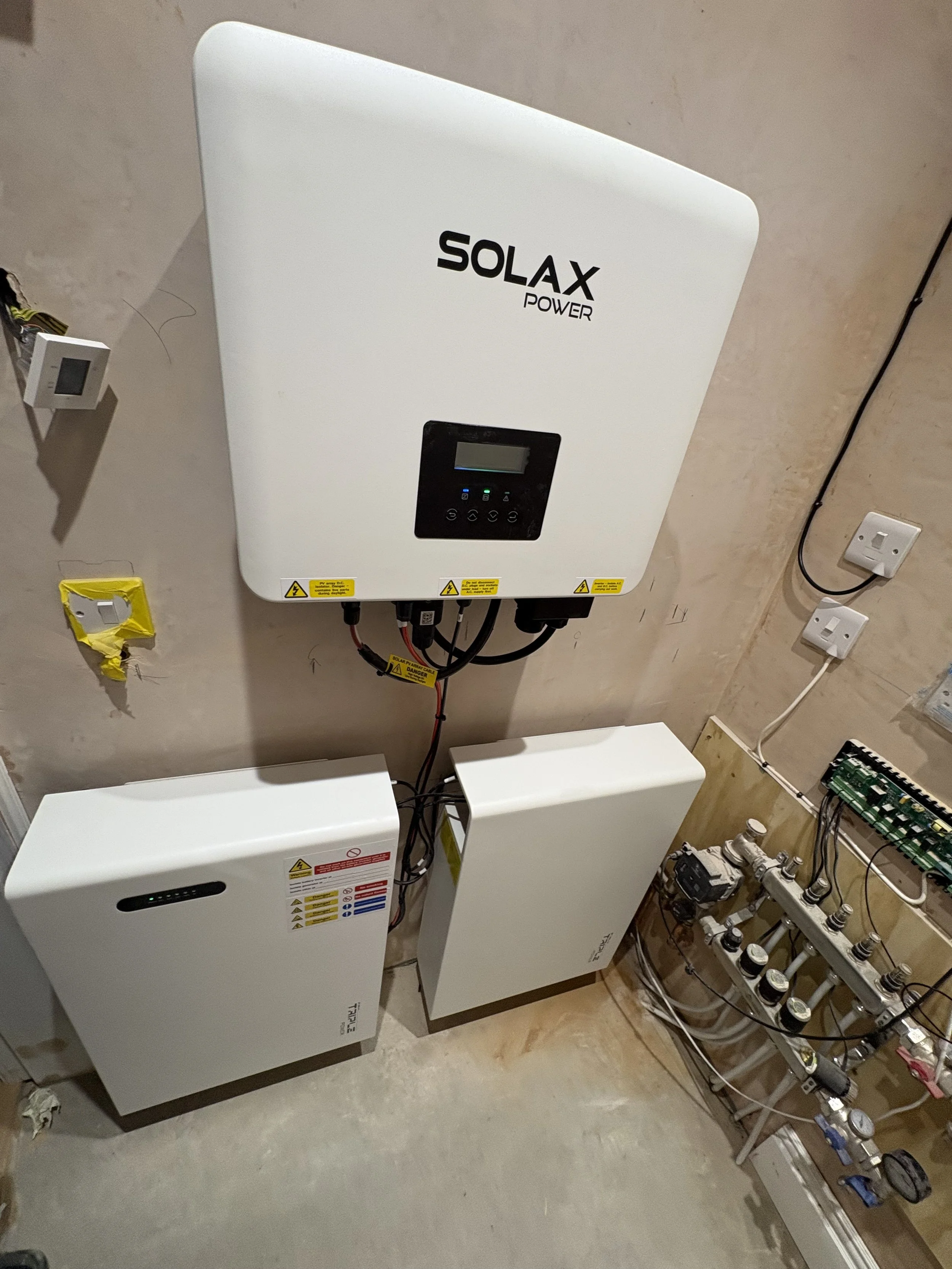 An electrical setup on a wall with a large white solar power inverter labeled 'SOLAX POWER', a white battery storage unit, and various pipes and gauges for a solar energy system.