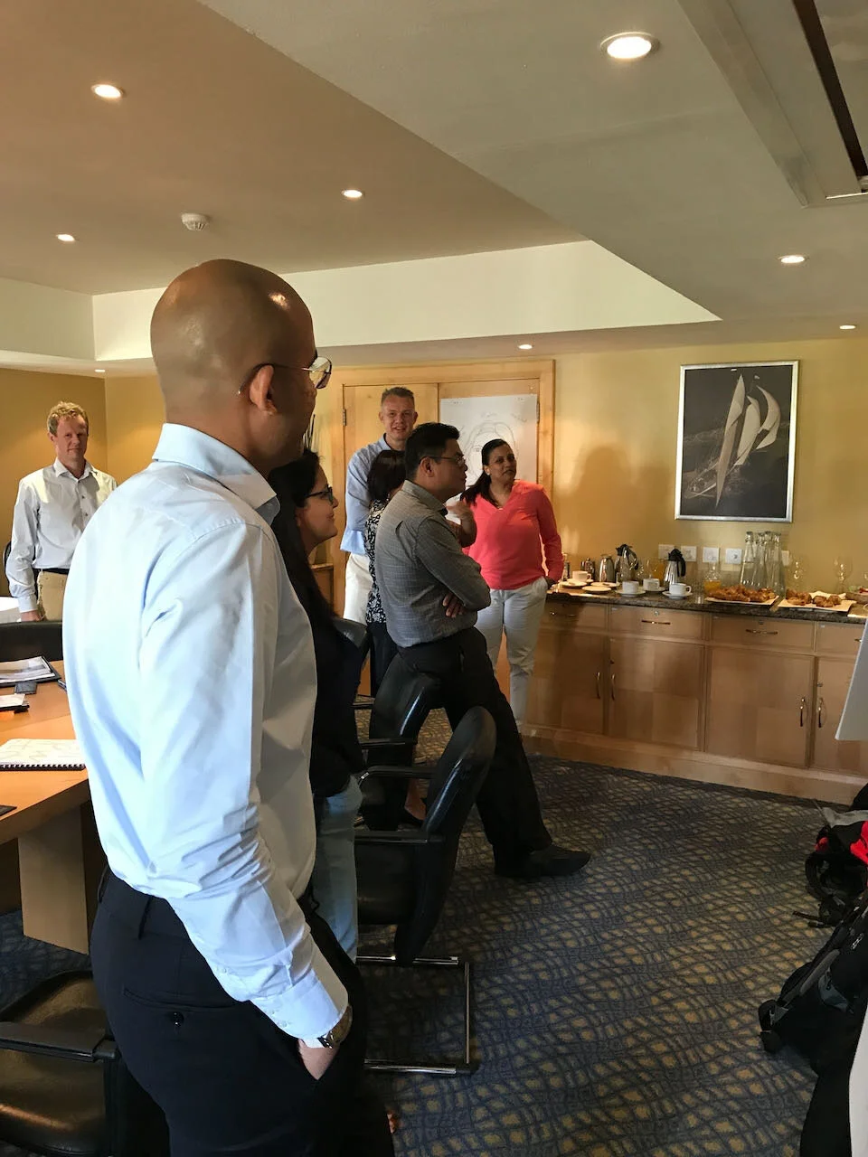 Group of professionals standing and listening in a conference room with a buffet table in the background.