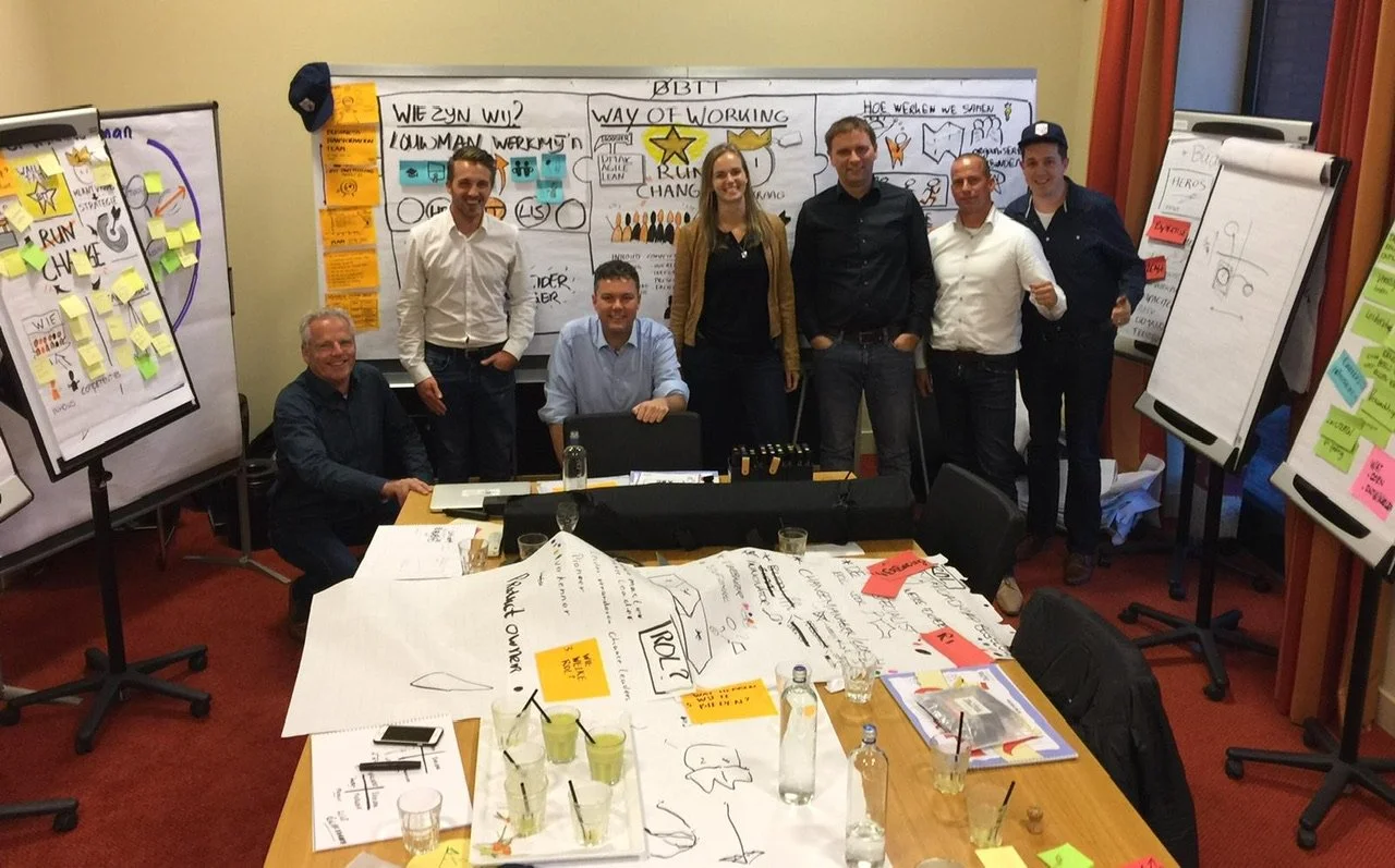 Group of seven people in a meeting or workshop room with whiteboards, flip charts, and tables covered with papers, sticky notes, and drinks.