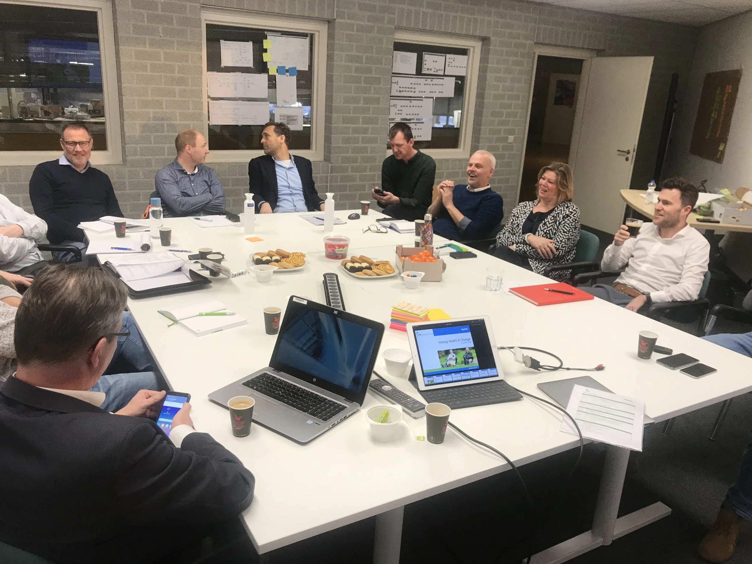 A group of people gathered around a conference table with snacks, drinks, laptops, and documents, engaged in a meeting or discussion in an office setting.