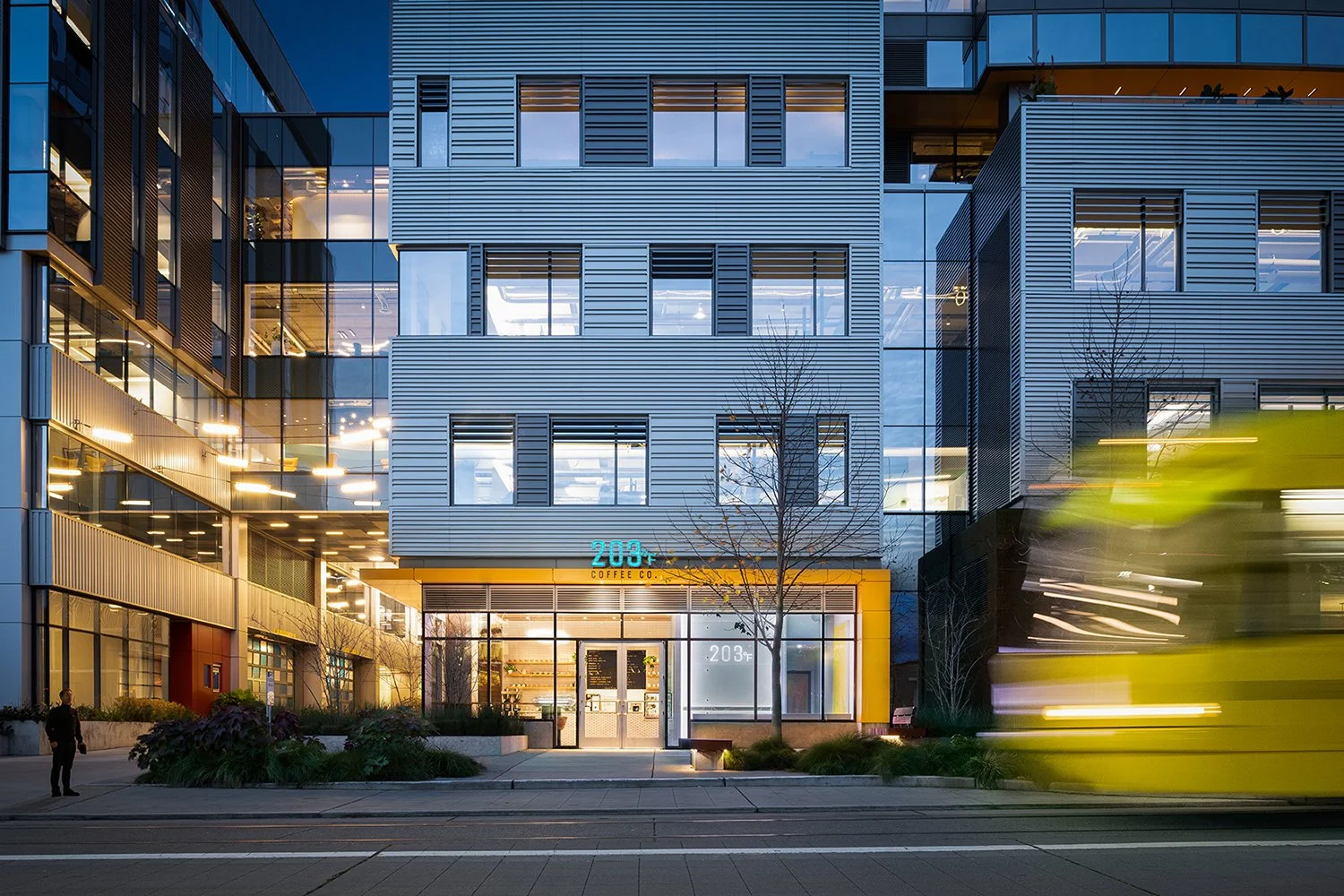 Modern building with glass and metal facade, featuring "203 Coffee Co." entrance, illuminated at night with a blurred yellow bus passing by.