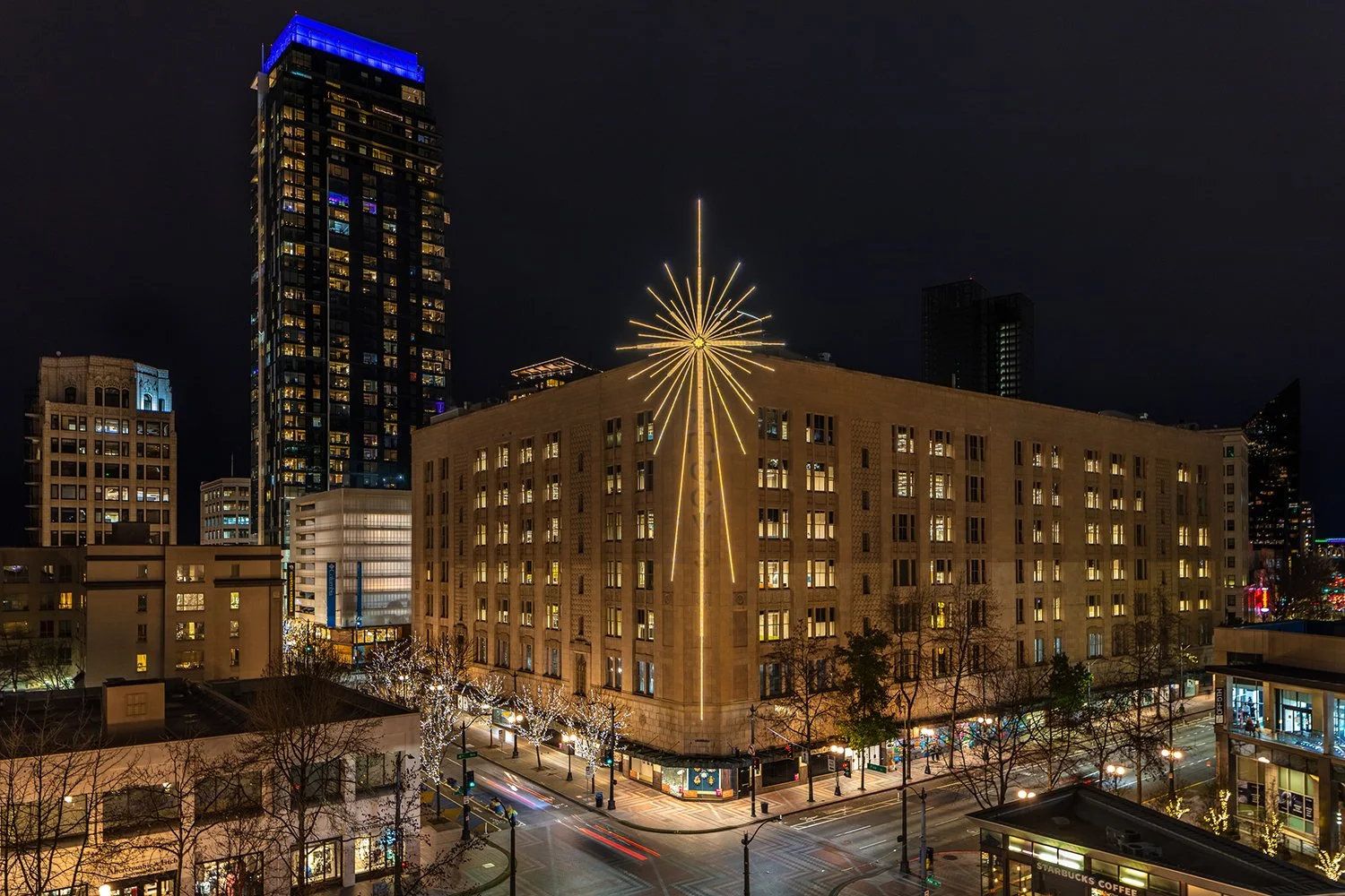 Cityscape at night featuring a tall building with a blue-lit crown, a large beige building with an illuminated decorative star, and streets lined with lit trees.