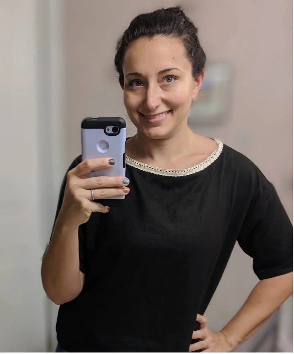 A woman with dark hair tied back, wearing a black shirt with a lace neckline, taking a selfie in front of a mirror. She is smiling and holding a smartphone with her right hand while her left hand rests on her hip.