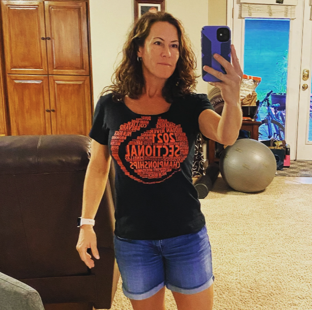 A woman taking a mirror selfie indoors wearing a black 'SEO NATIONAL' T-shirt and blue denim shorts.
