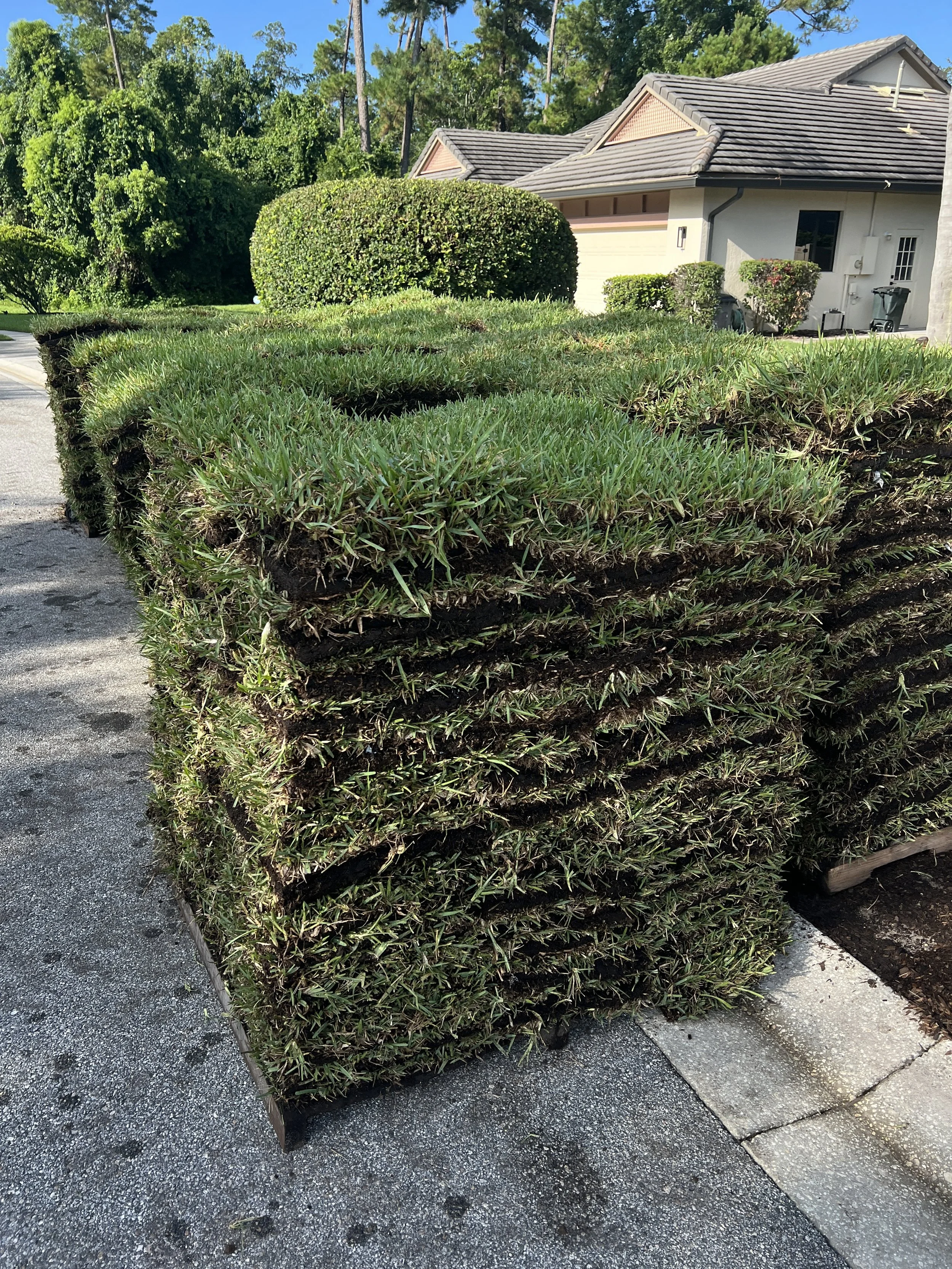 Multiple stacked bundles of freshly cut grass or sod outdoors near a house with a garage, trees, and shrubs in the background.