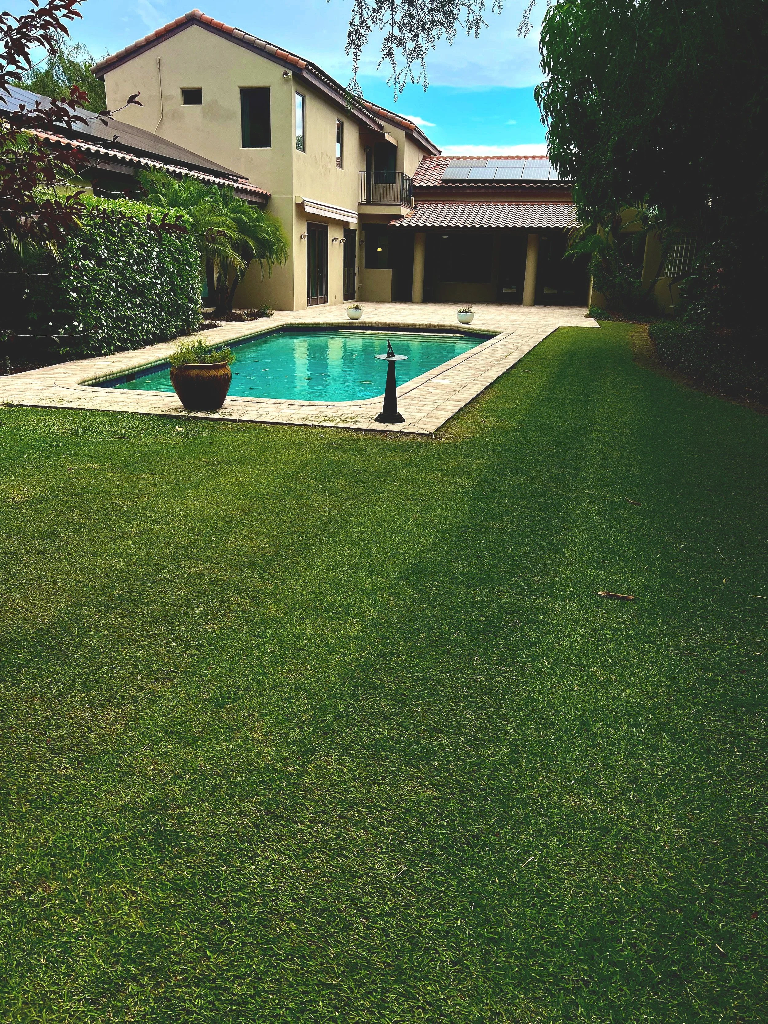Backyard with a swimming pool, lush green lawn, and a beige house with a red-tiled roof under a partly cloudy sky.