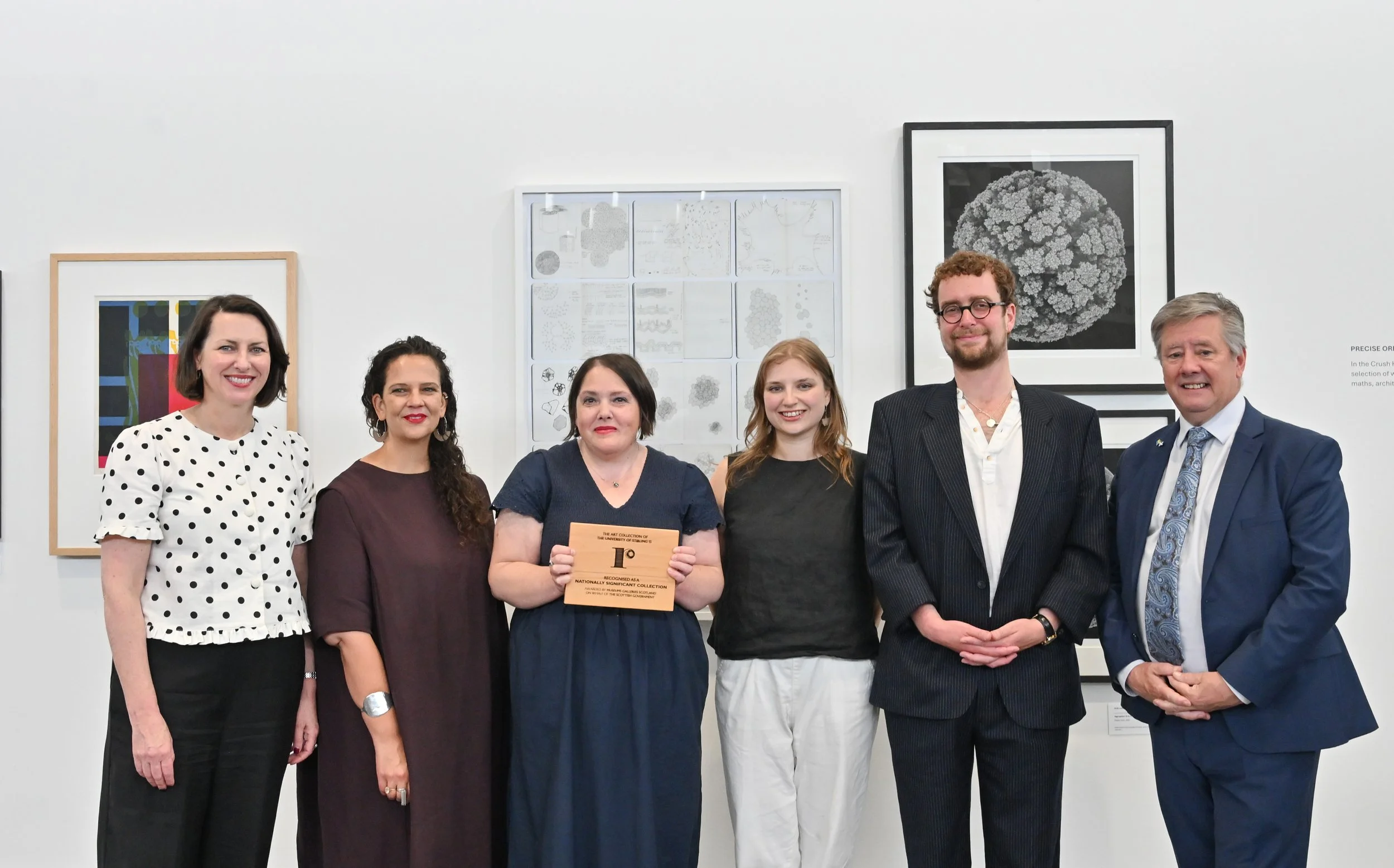 Award ceremony photographed by julie Howden in the Crush Hall in the Pathfoot Building in the university of Stirling with the art collection behind. four women stand in a row with two men. the third woman is holding a plaque from wood with black text