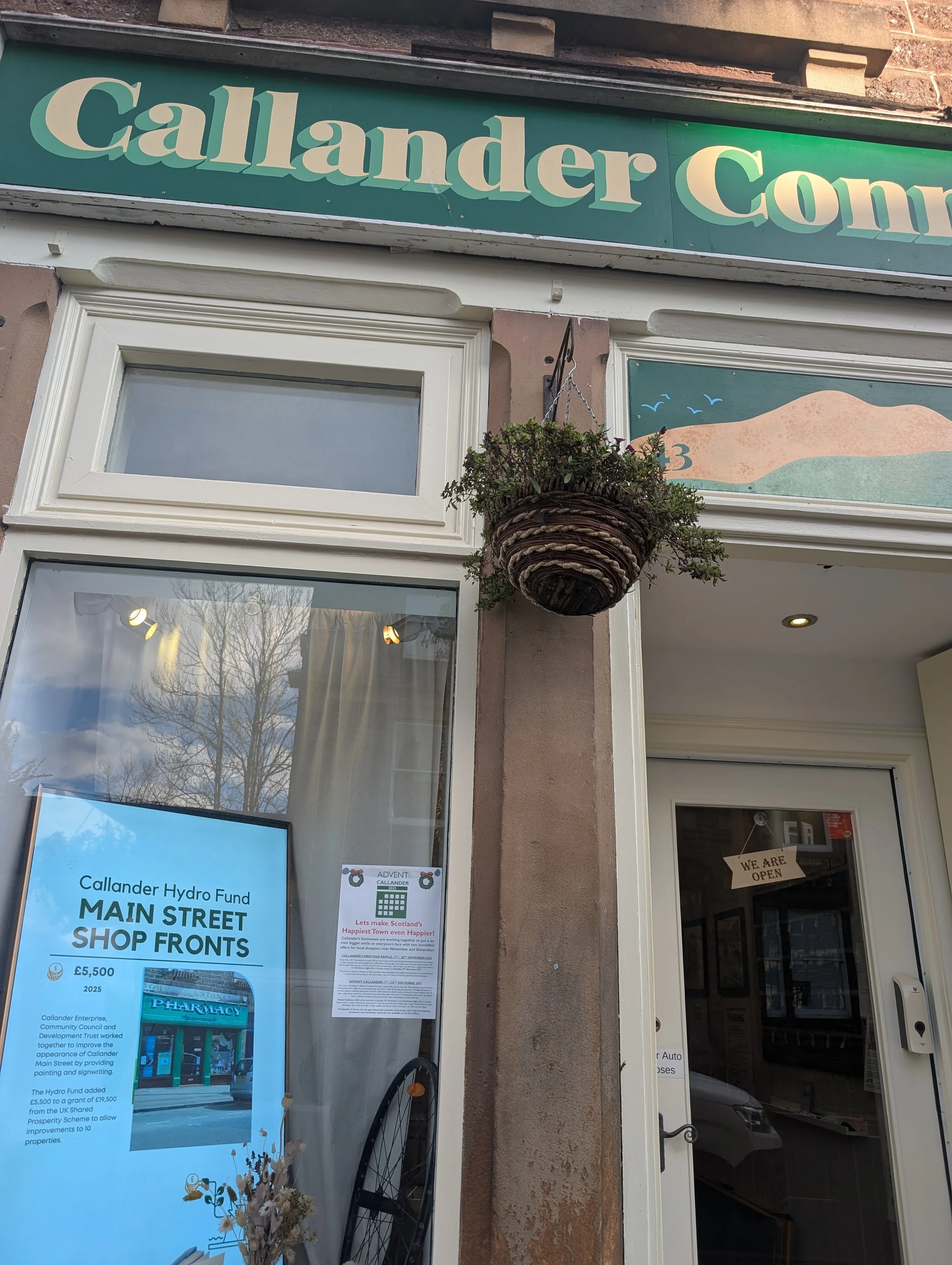 The Callander Connect shopfront on Main Street, Callander, with a green painted sign, hanging flower basket, and a window display about the Callander Hydro Fund's Main Street Shop Fronts project.