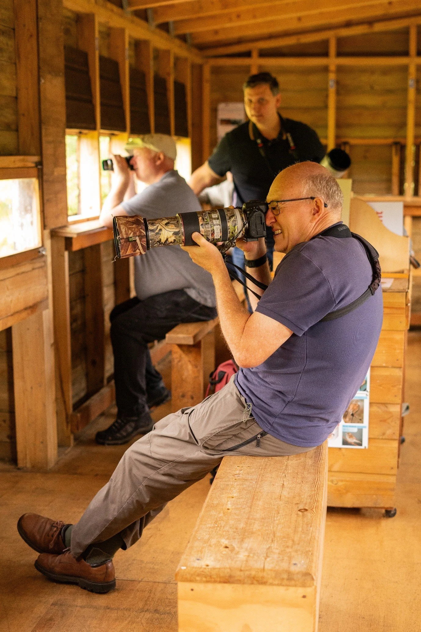 "Argaty Red Kites" (2025) by Anne Sproul, for Stirling Photography Festival — three people inside a wooden hide use long-lens cameras and binoculars to photograph wildlife through the hide's open windows.
