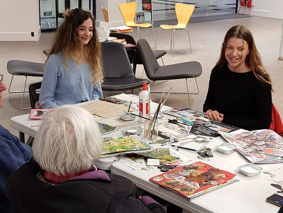 An intergenerational collage-making workshop, with participants of different ages sitting around a table covered in cut-out images, glue and paintbrushes, working on collage artwork together.