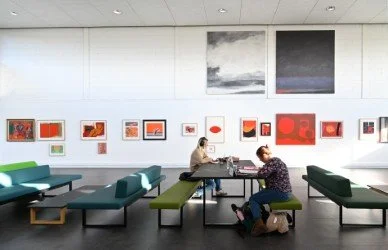 Two people sitting at a table in the Crush Hall of the Pathfoot Building, University of Stirling, home to the University's Art Collection, with artworks displayed on the walls and green bench seating in the foreground.