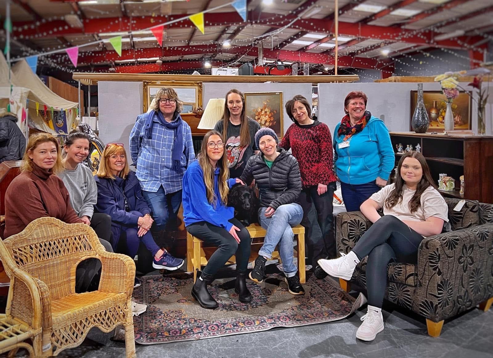 A group of staff and volunteers, plus a dog, sitting together in a lounge-style seating area at the Stirling Reuse Hub, surrounded by donated furniture and decorative bunting.
