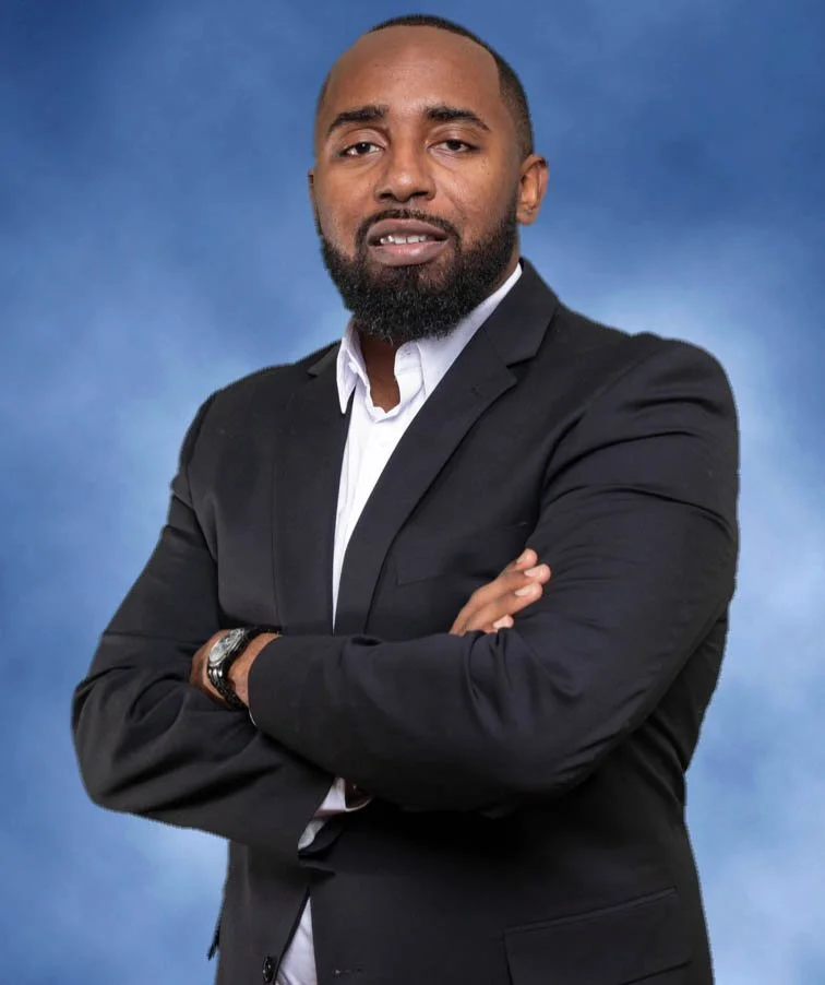 A man with a beard and short hair, dressed in a black suit and white shirt, standing with arms crossed against a blue background.