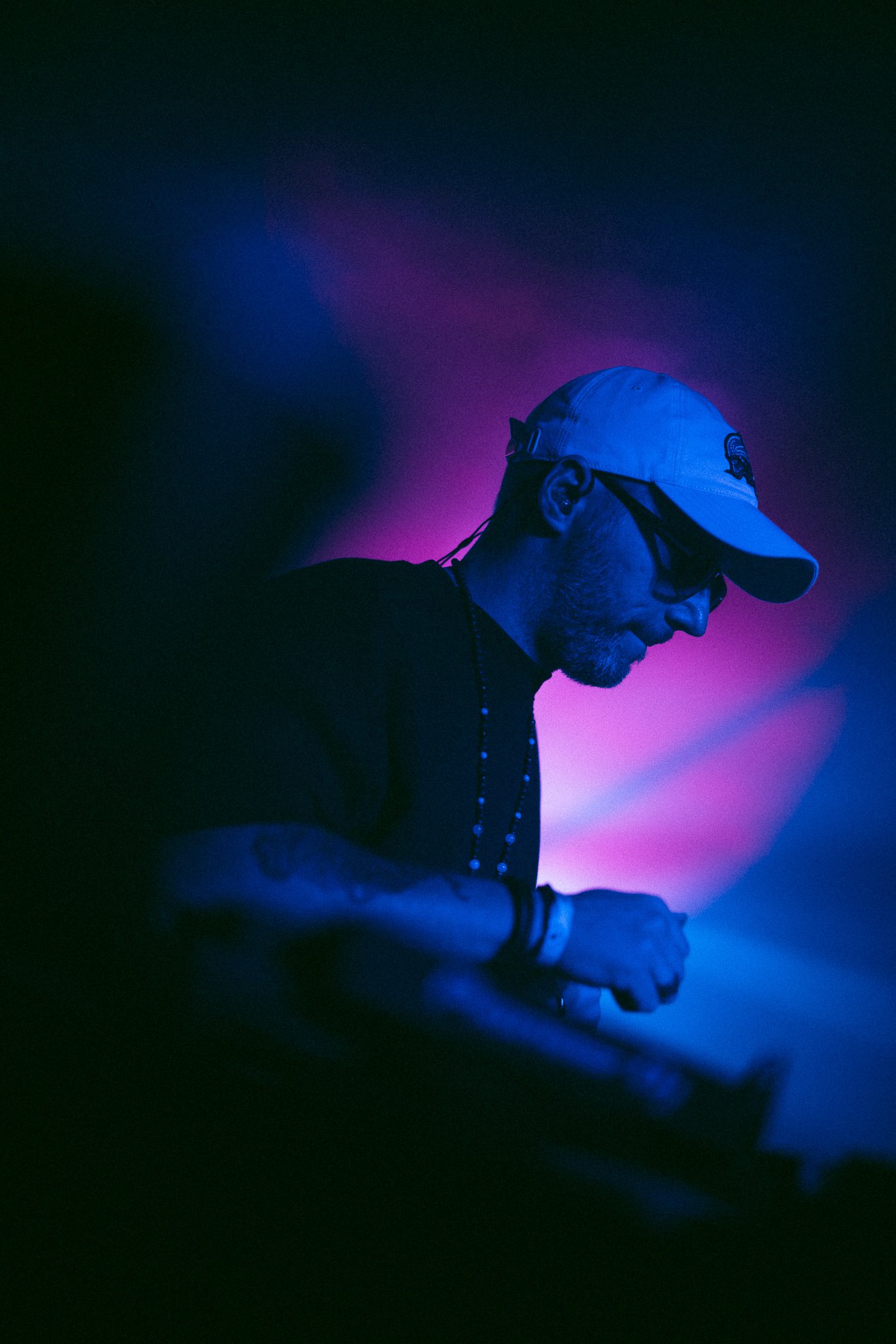 A male DJ wearing a baseball cap and glasses, illuminated with purple and blue lighting, working at a DJ booth