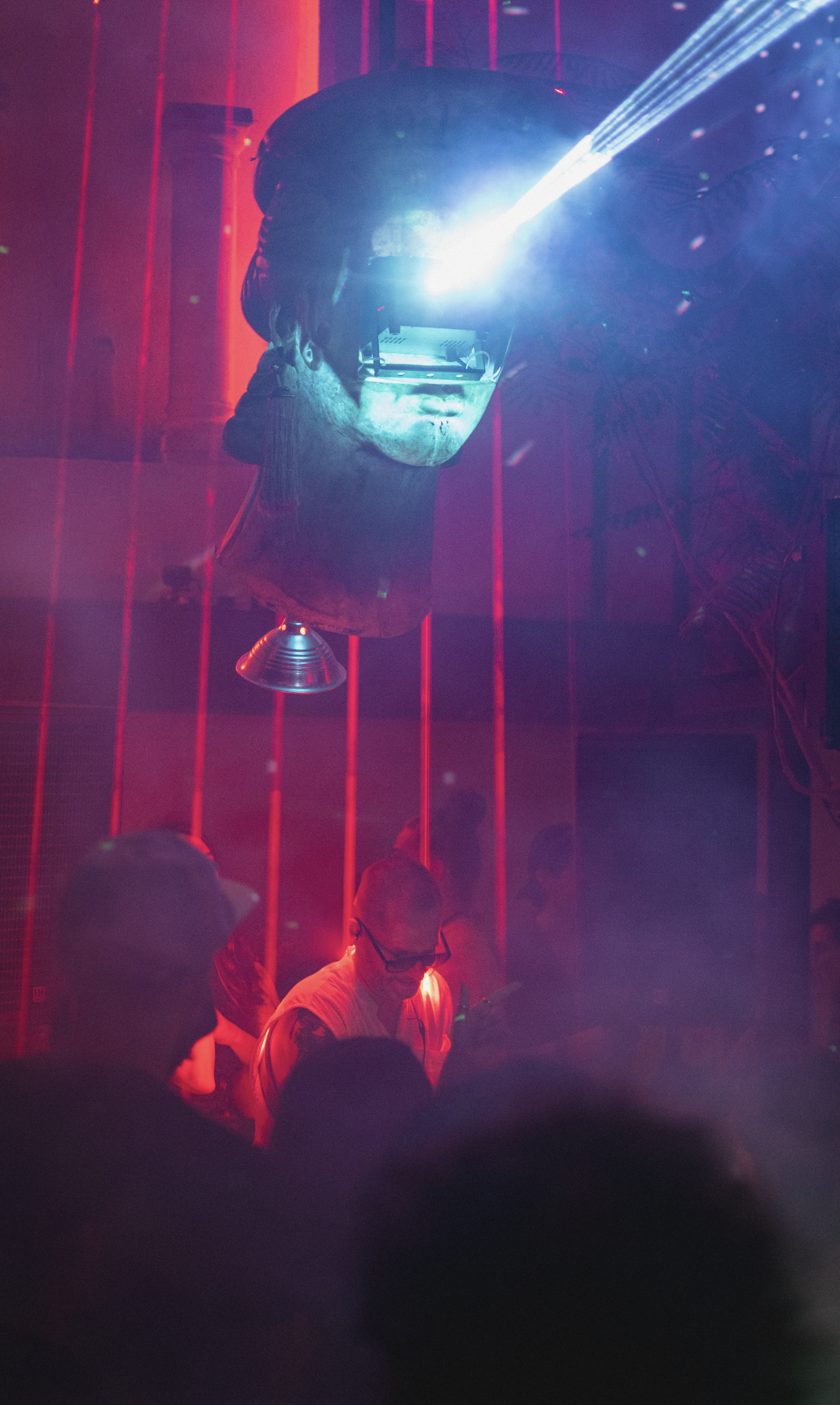 A digital art installation features a large human head sculpture wearing a virtual reality headset with bright blue light emerging from it, set in a dark room with red vertical light bars and faint crowd silhouettes.