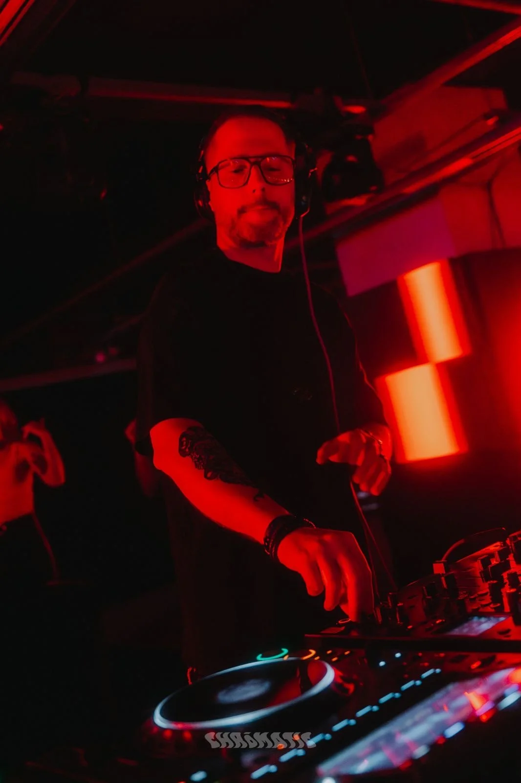 A DJ wearing glasses, headphones, and a black shirt, standing behind a DJ console in a dimly lit club with red lighting.