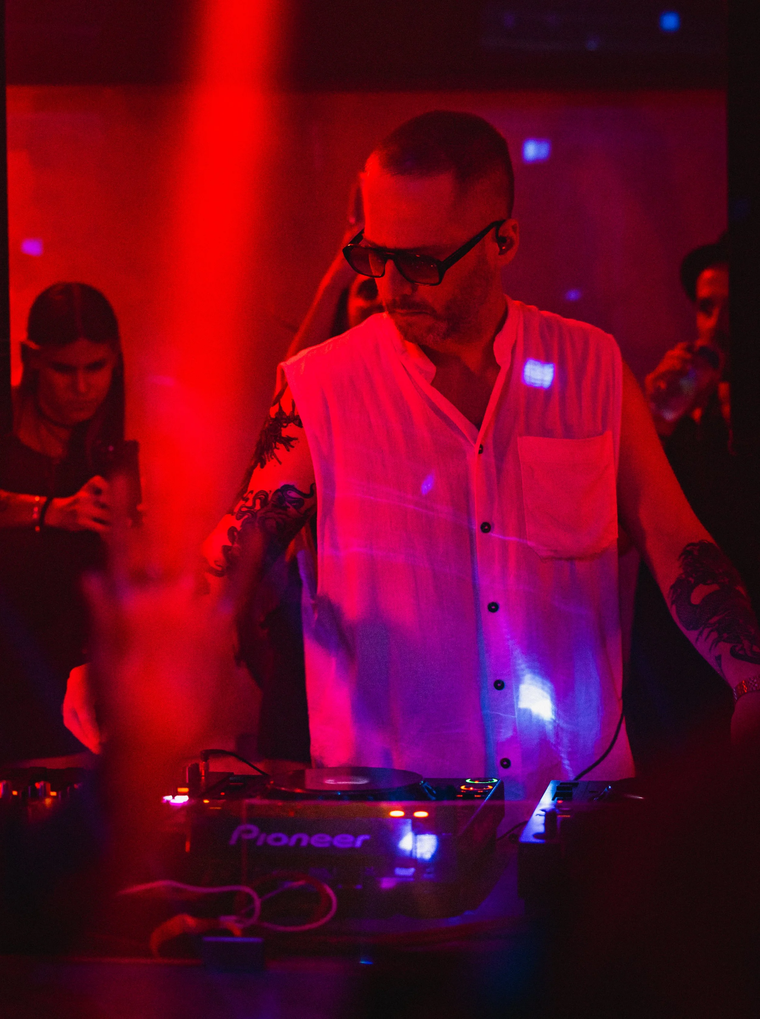 A man with tattoos on his arms, wearing sunglasses and a sleeveless white shirt, is DJing at a nightclub with red lighting, surrounded by people, some of whom are holding drinks.