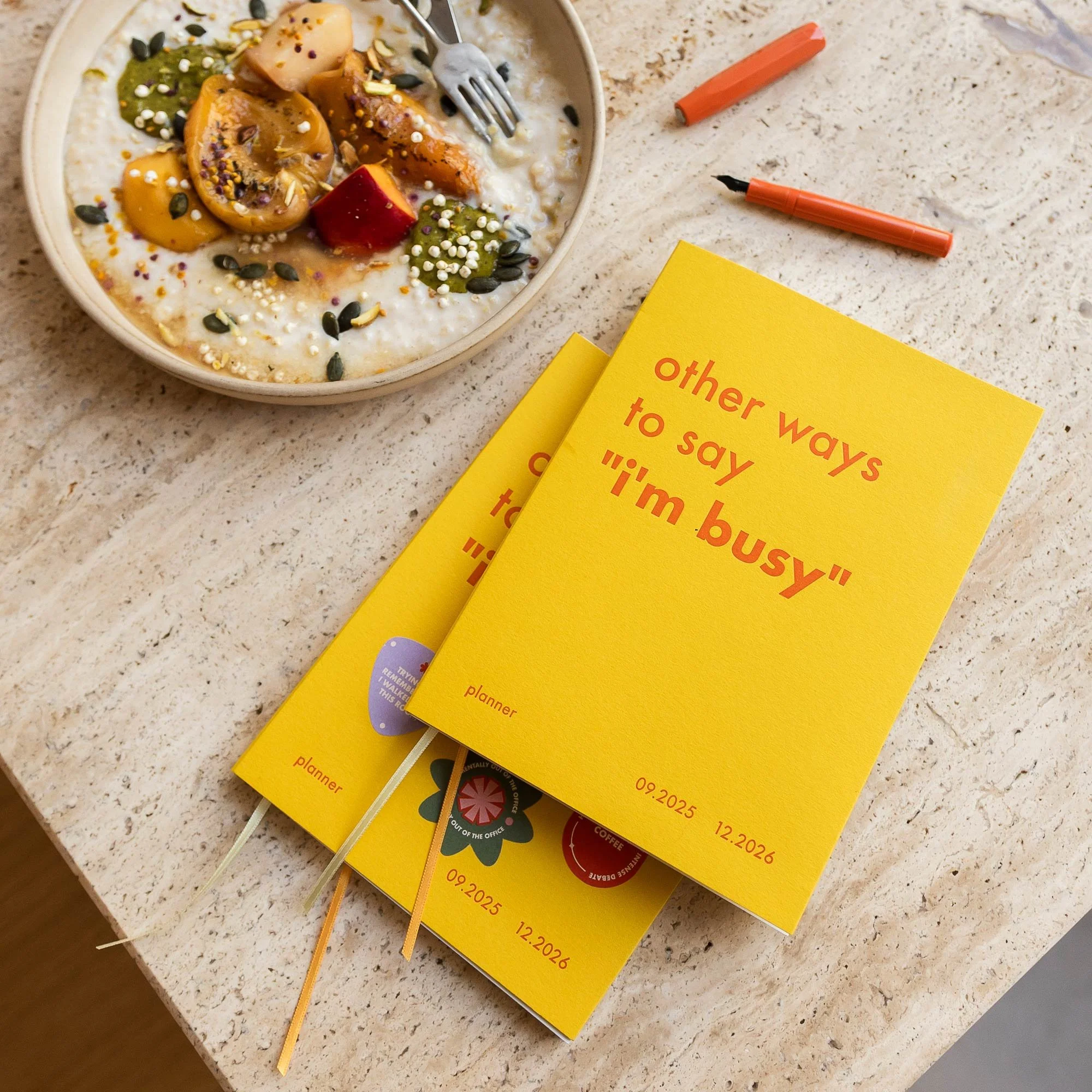 Two books standing on a marble surface, one beige and one blue, both with a textured cover and a date range including 2023; a framed abstract art piece in the background.