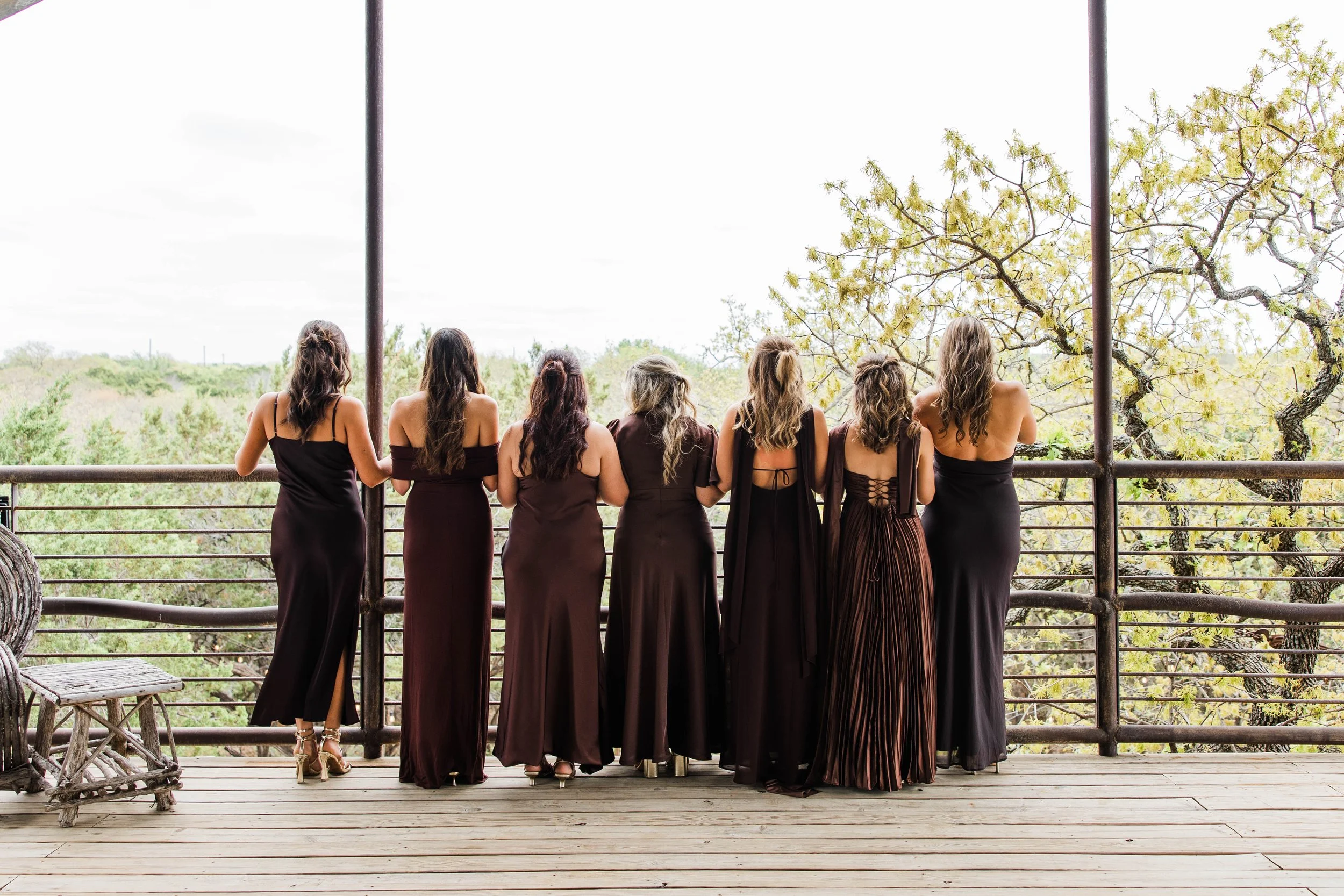 Bridesmaids looking off the balcony at Sparrow Creek Ranch. They are all wearing chocolate brown bridesmaids dresses looking beautiful against the tops of green oak trees.