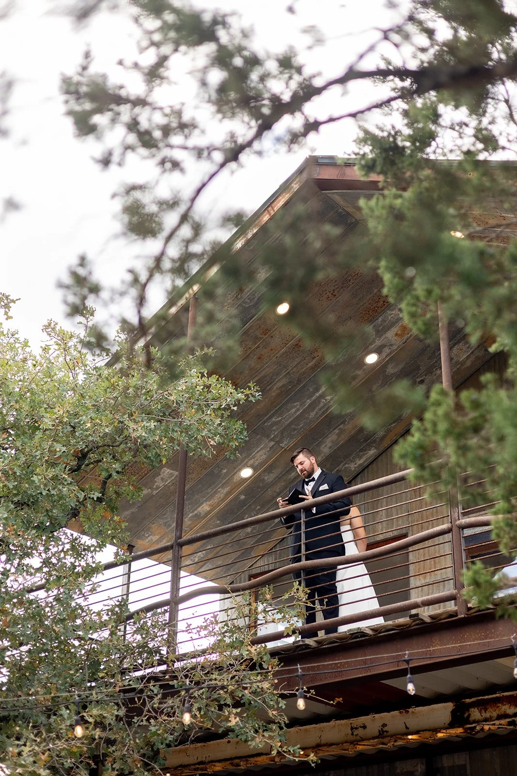 groom standing on the balcony surrounded by trees with the bride turned so they are back to back for a private pre-ceremony vow exchange at Sparrow Creek Ranch wedding venue.