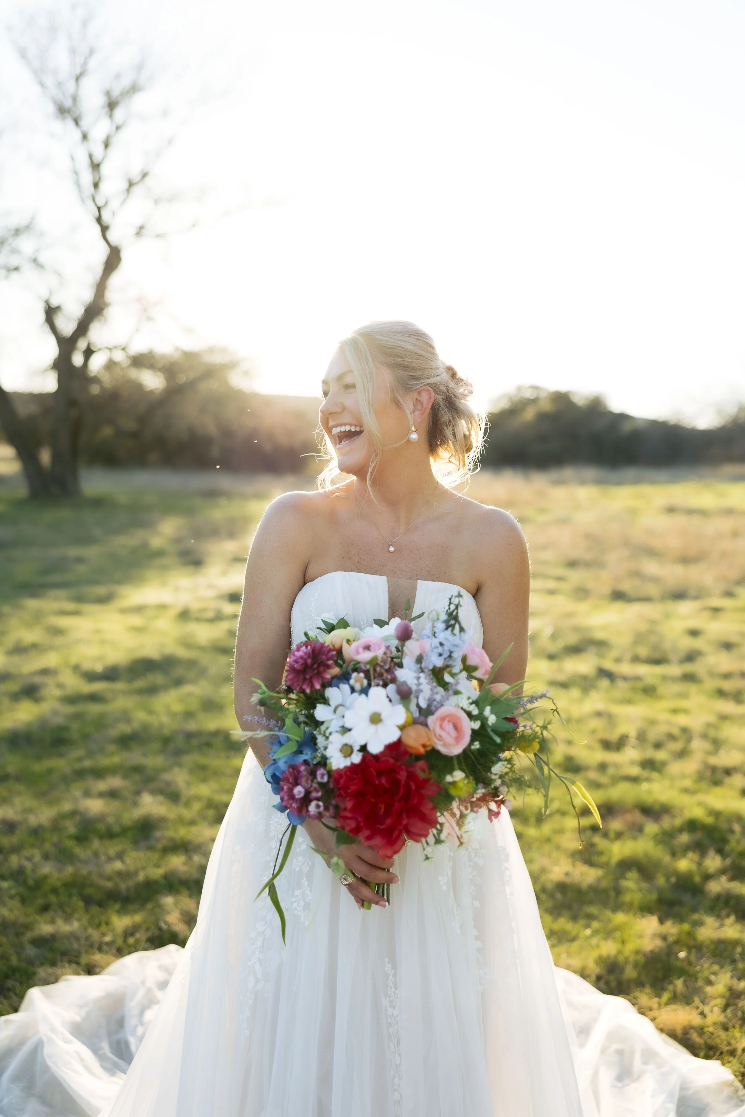 Bride laughs while taking bridal session photos holding a bibrant spring bouquet of white pink and light blues with one large red color flower for a big pop.
