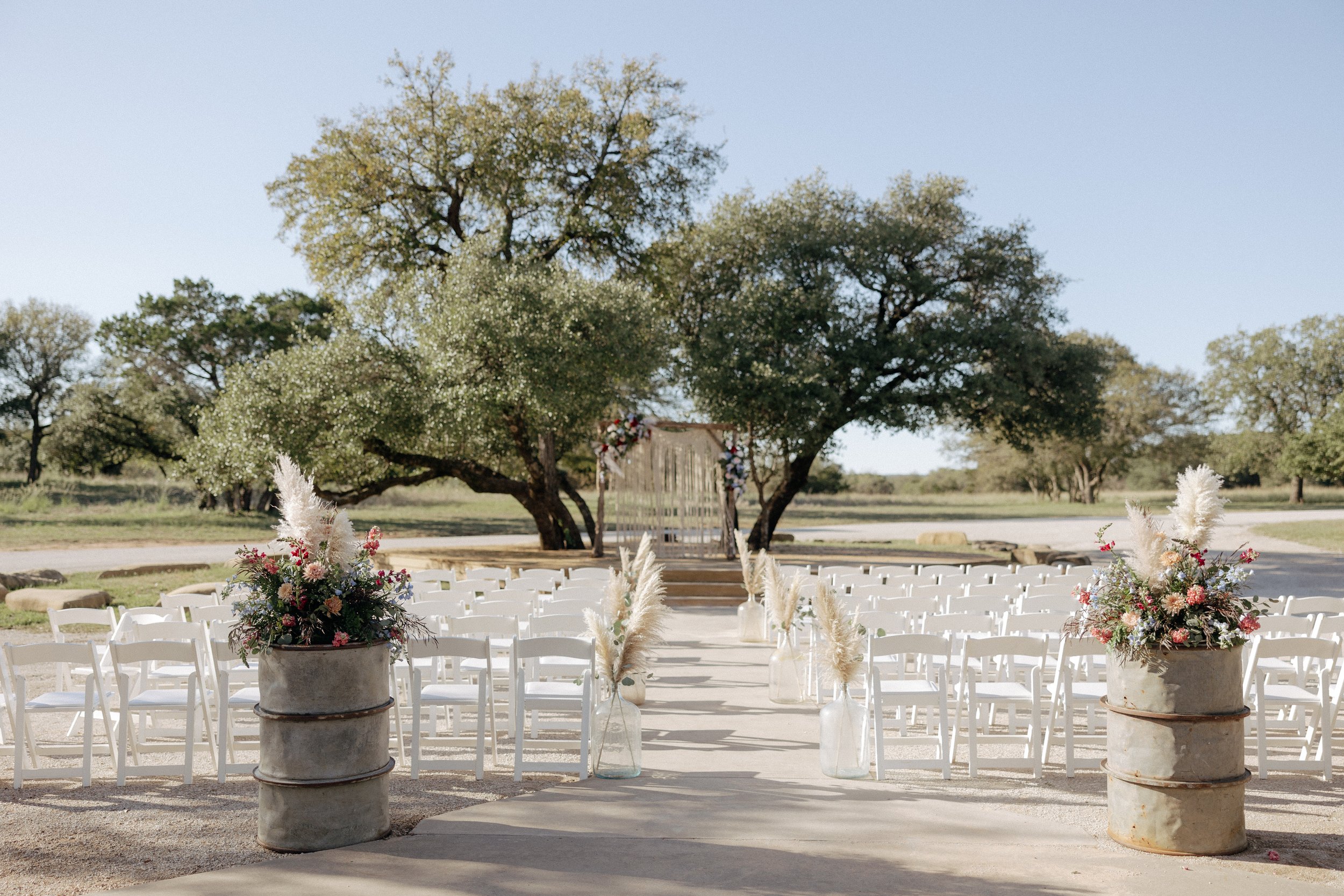 November wedding outside at the ranch venue near dfw named Sparrow Creek Ranch. This ceremony setup has two old oil barrels for forals, white garden chairs, and big oak trees.