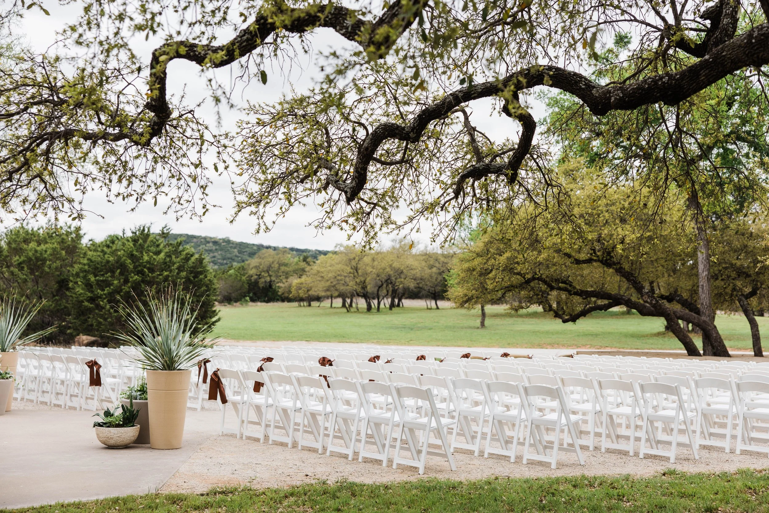 Stunning outdoor ceremony setting on a ranch in North Texas with big oak trees surrounding the chairs and platform. The landscape is rolling hills all in new spring growth at the end of March.