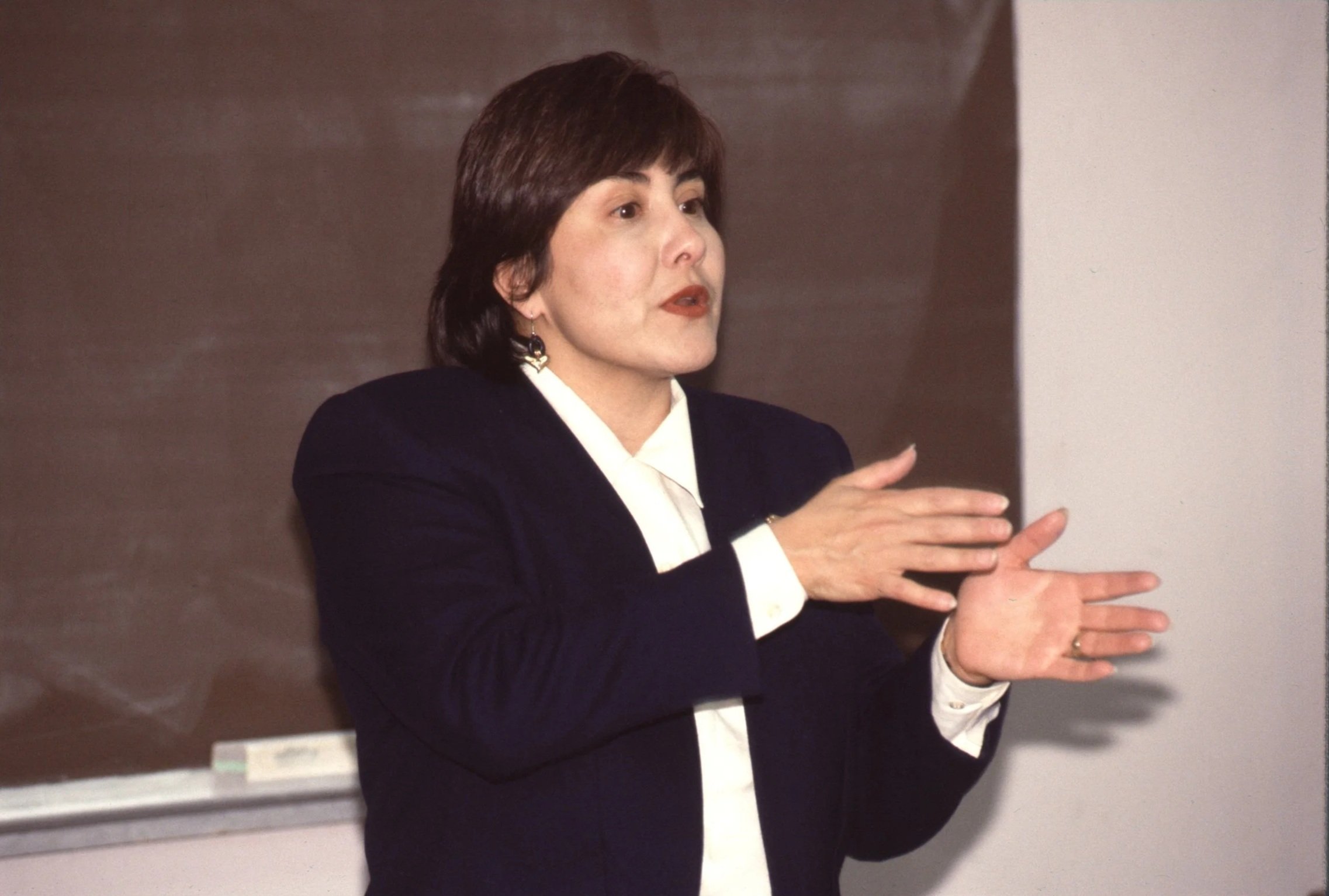 A woman with short dark hair, wearing a black blazer and white blouse, gesturing with her hands while speaking in front of a dark and light-colored wall.
