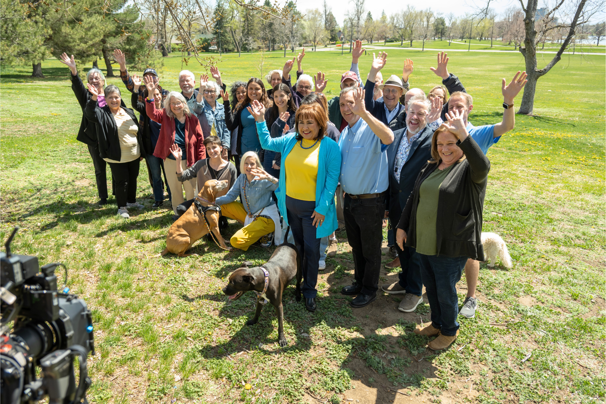 A large group of smiling diverse people standing outdoors in a park on a sunny day, waving at a camera for a group photo, with some dogs present.