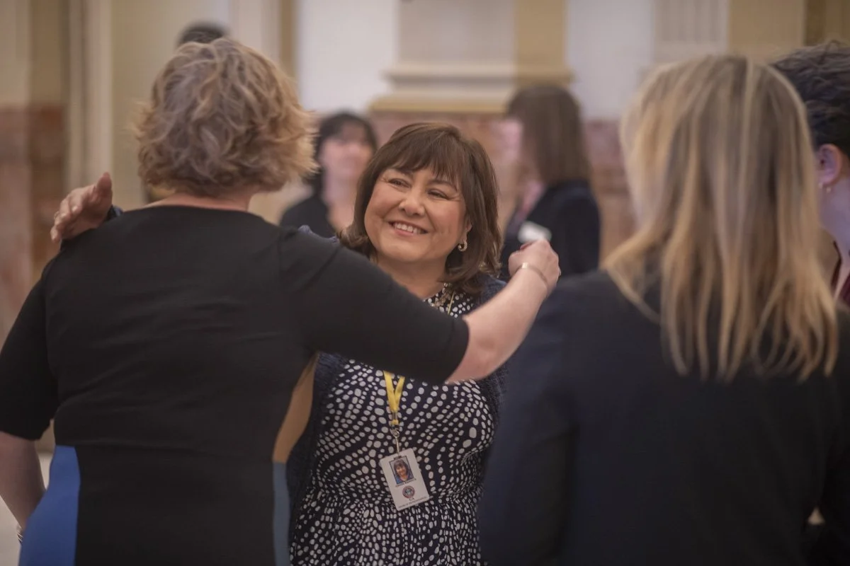 A woman smiling while hugging another woman at a social gathering or conference, surrounded by other women engaged in conversation.