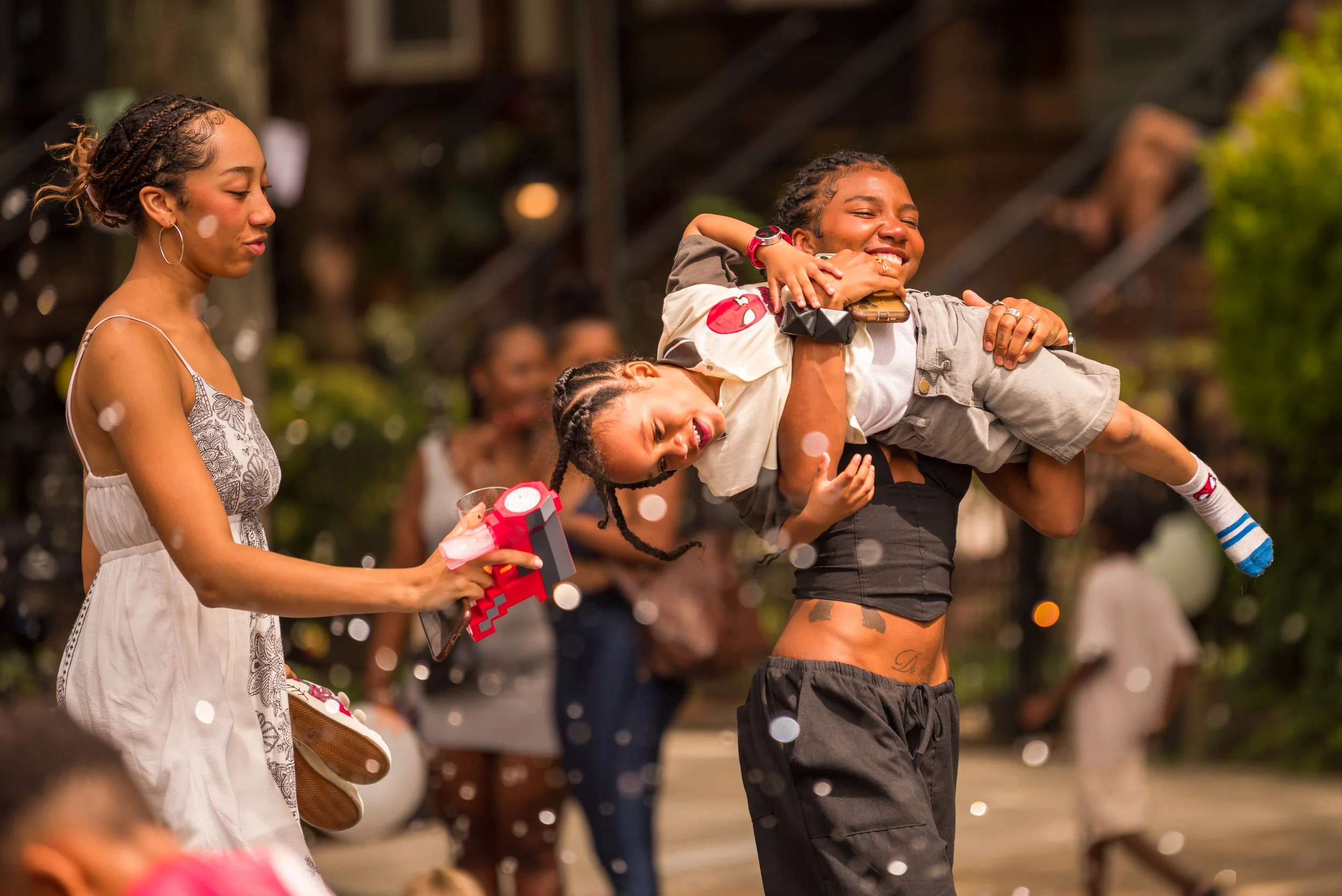 A woman with braids holding two children, one over her shoulder and the other in her arm, smiling at a celebration or outdoor gathering. The woman is wearing black shorts, and the children are in casual clothes. Confetti is falling, and people are visible in the background.