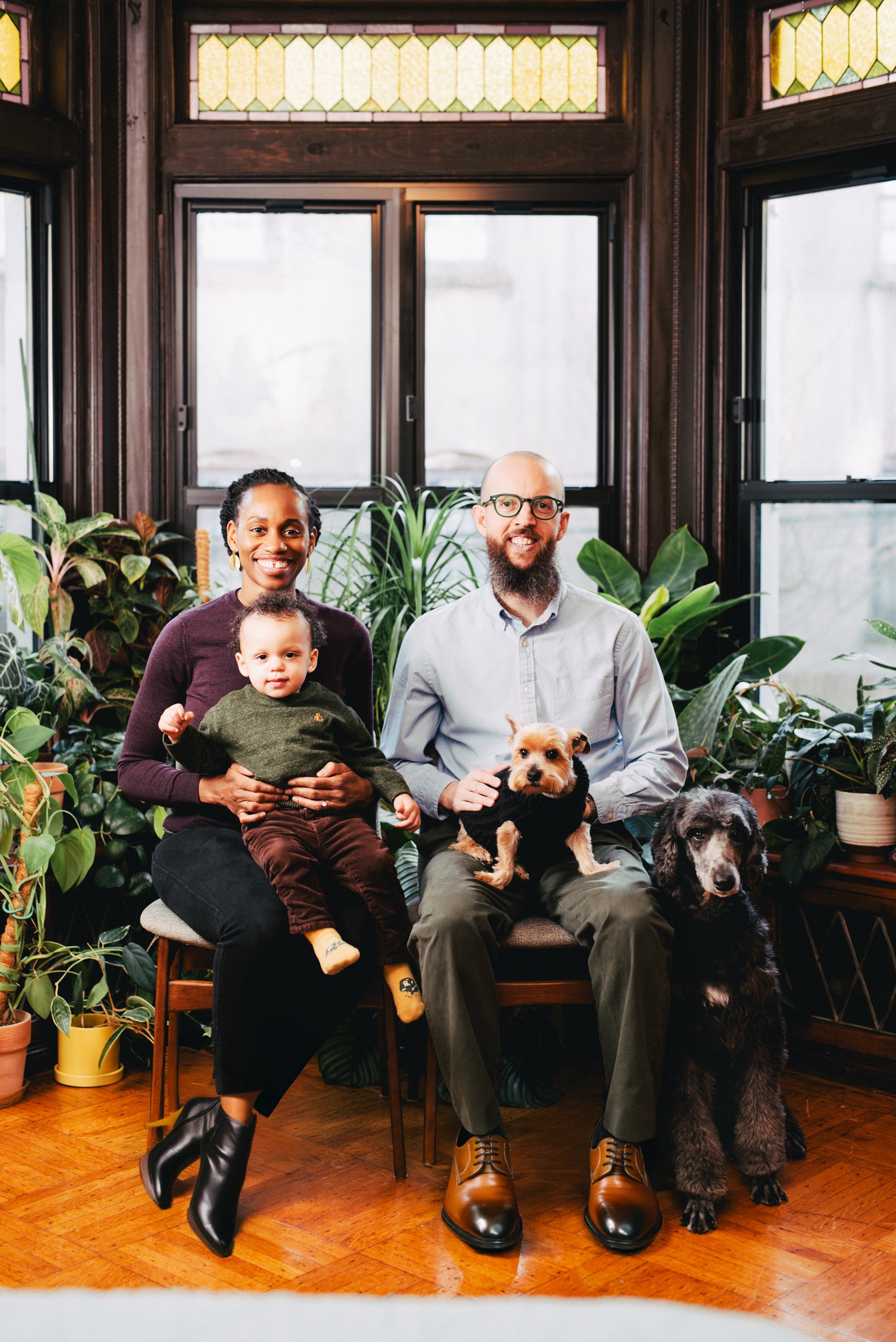A family of four, including two dogs and a young child, sitting together in a cozy room with large windows and surrounded by indoor plants.
